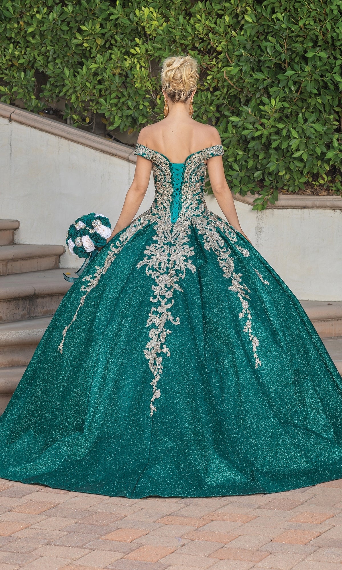 A woman stands outdoors, facing away from the camera, wearing Quinceanera Dress 1753 By Dancing Queen—an emerald green off-the-shoulder ball gown with silver lace embroidery—while holding a bouquet.