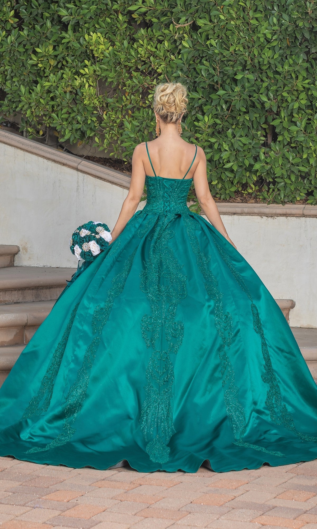 A woman in the Quinceanera Dress 1756 By Dancing Queen, a teal a-line ball gown with lace embroidery, stands on steps holding a bouquet, facing away amid lush green foliage.