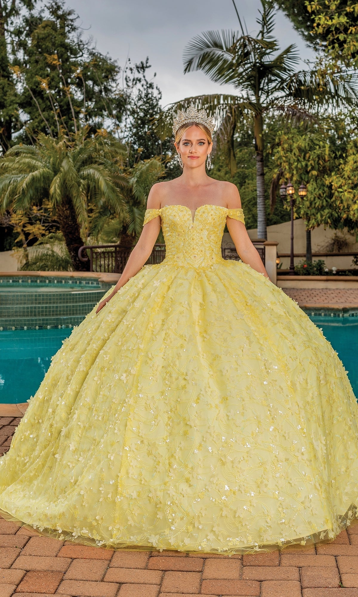A woman stands outdoors by a pool, wearing the Quinceanera Dress 1757 By Dancing Queen—a yellow ball gown with off-the-shoulder straps and floral embroidery—paired with a tiara and surrounded by lush greenery.