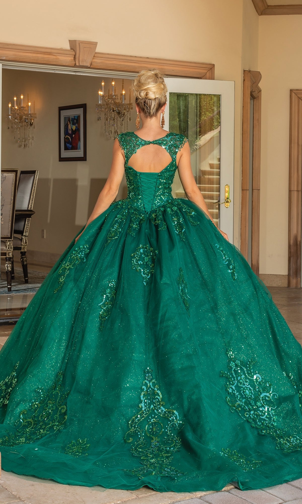 A woman with blonde hair in an updo wears the Quinceanera Dress 1758 by Dancing Queen, a green embellished a-line ball gown with glitter lace embroidery, cutout back, and cap sleeves as she stands indoors facing away from the camera.