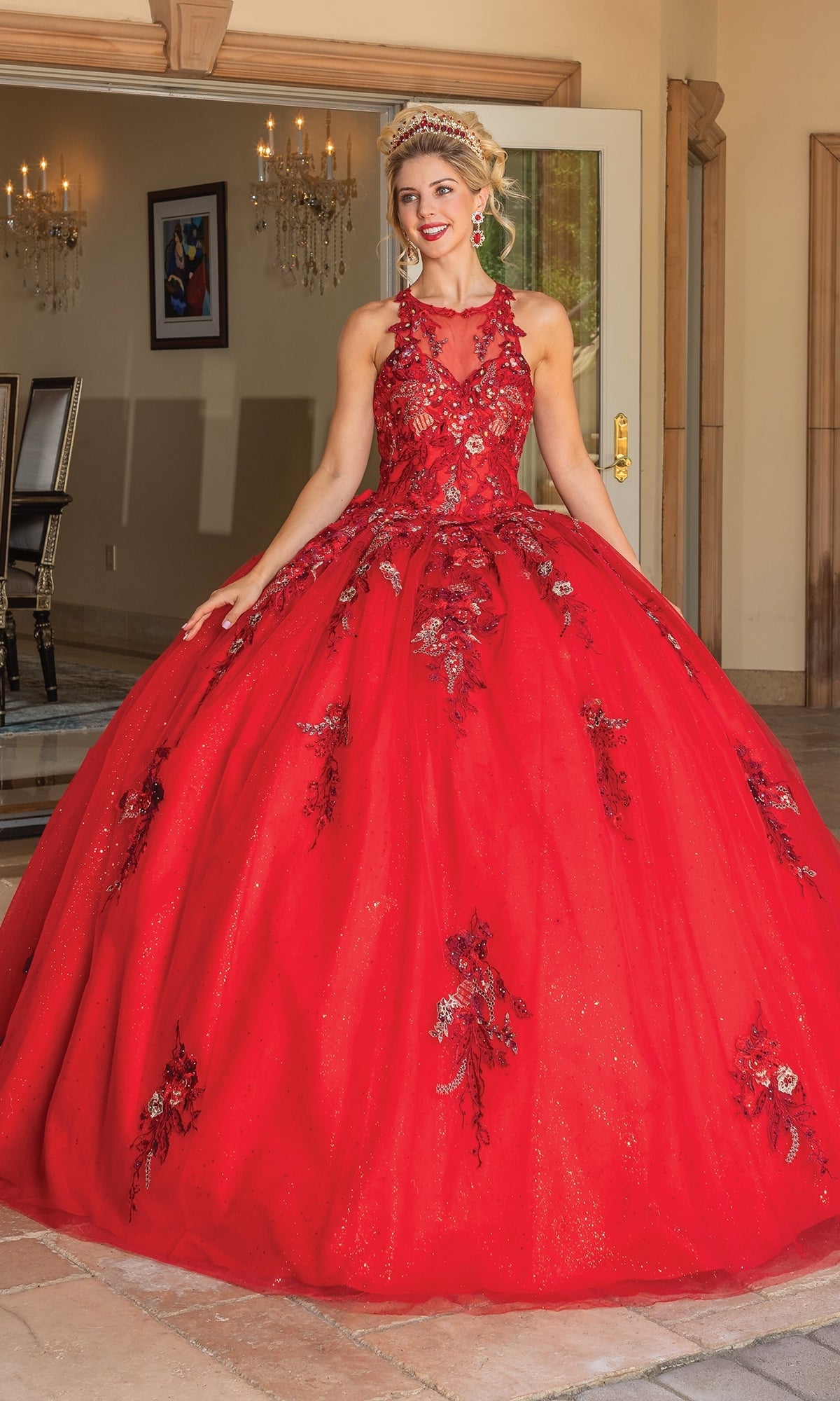 A woman smiles indoors near an open door, wearing the Quinceanera Dress 1761 By Dancing Queen—an ornate, bright red gown with floral embroidery and a dramatic bow back.