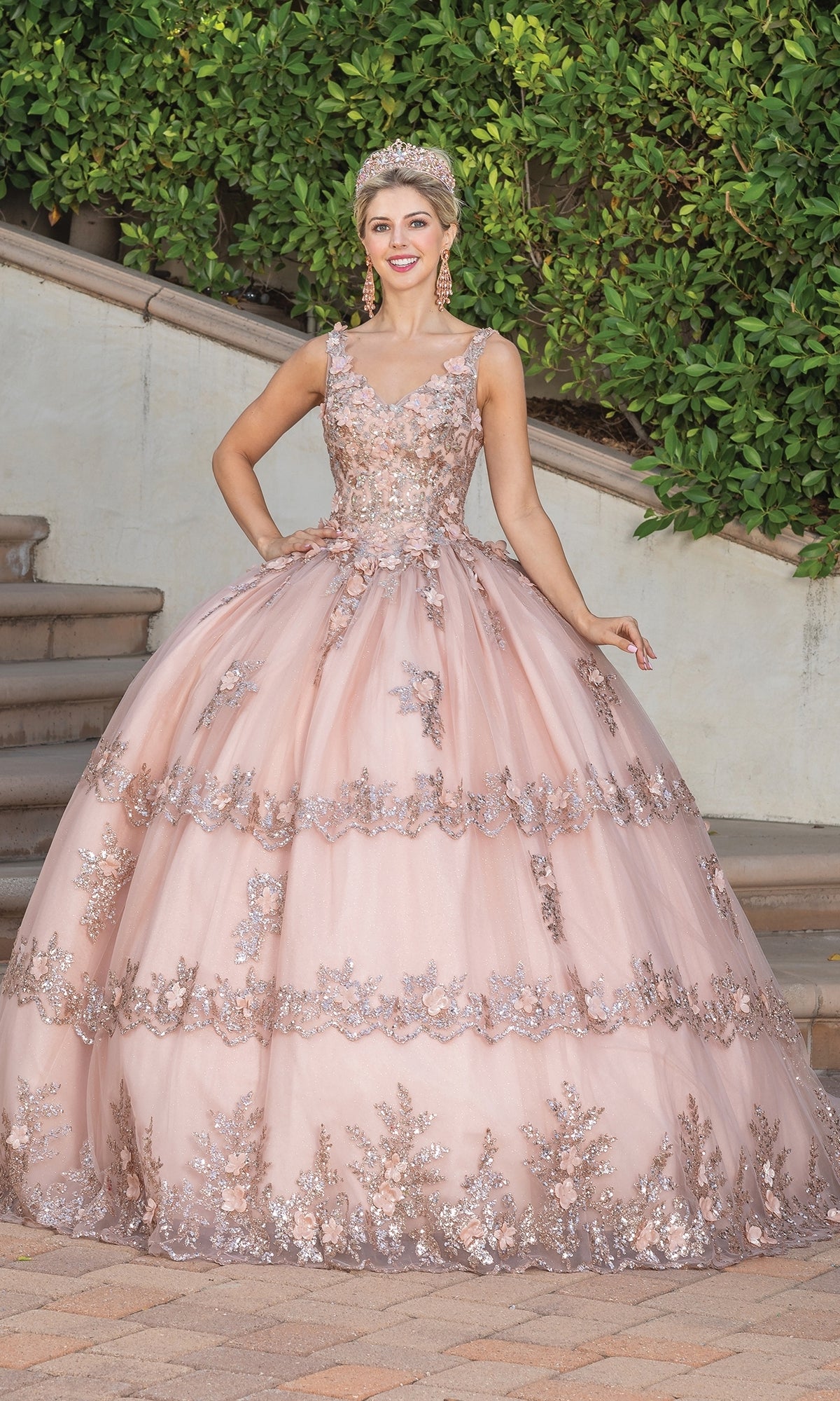 A young woman poses outdoors in front of steps and greenery wearing the Quinceanera Dress 1763 By Dancing Queen, an a-line ball gown featuring floral lace detailing and delicate applique.