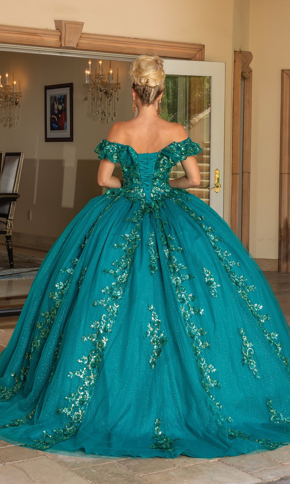 A woman stands indoors facing away, wearing the Quinceanera Dress 1766 By Dancing Queen—an a-line teal ball gown with off-the-shoulder sleeves, floral embellishments, and a full skirt.