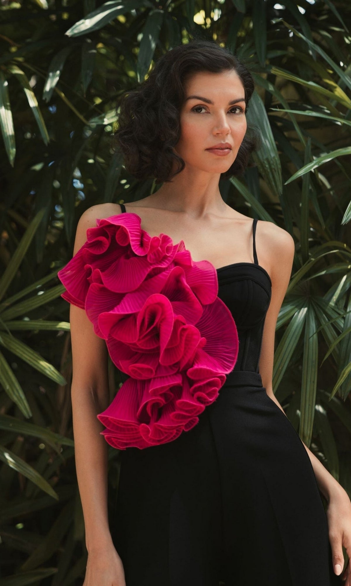 A woman stands before green foliage wearing the Long Formal Jumpsuit 11767 by Mac Duggal—black, with wide legs and a bold pink ruffle accent on one shoulder.