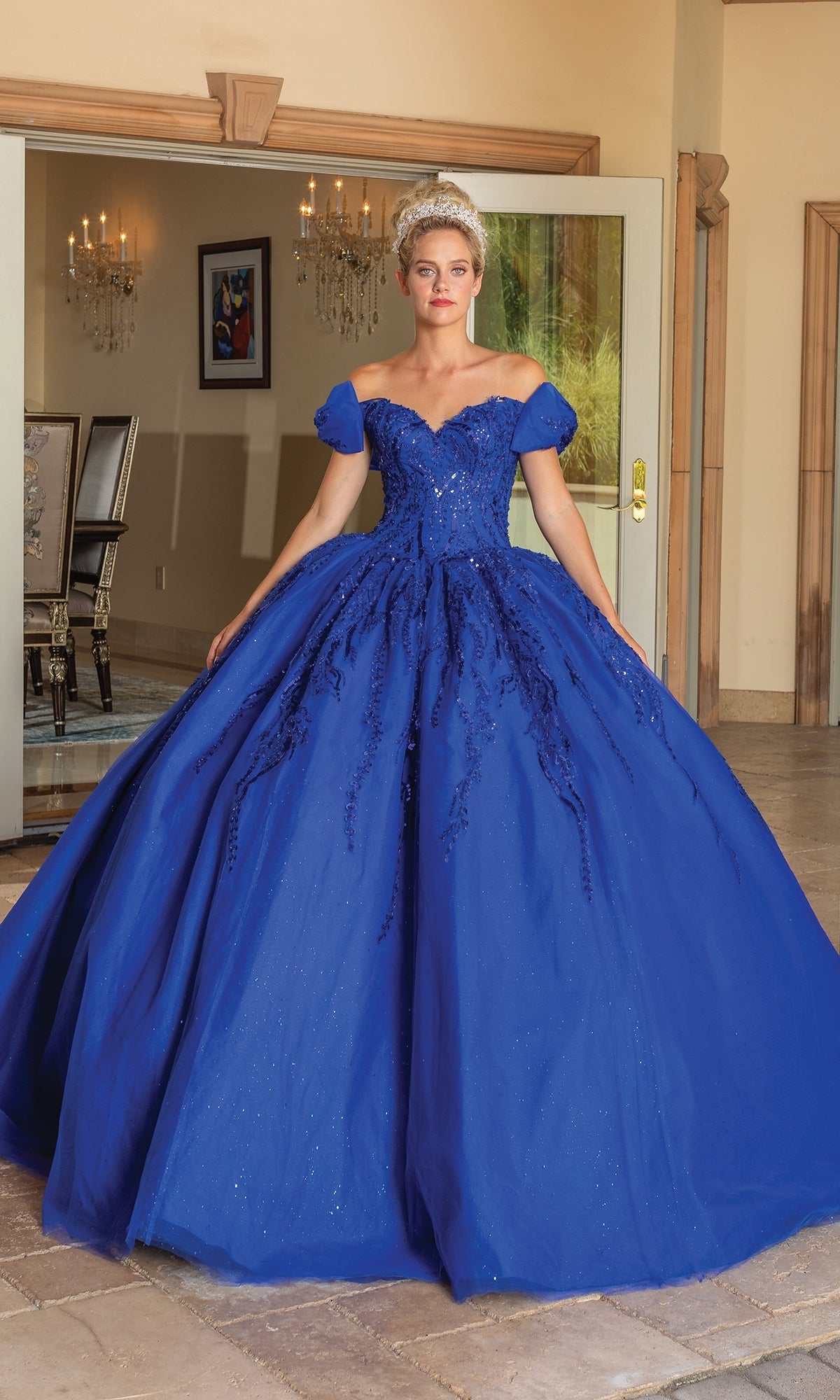 A woman stands indoors wearing the Quinceanera Dress 1780 By Dancing Queen, an elegant royal blue off-the-shoulder ball gown with intricate detailing and a voluminous full skirt.