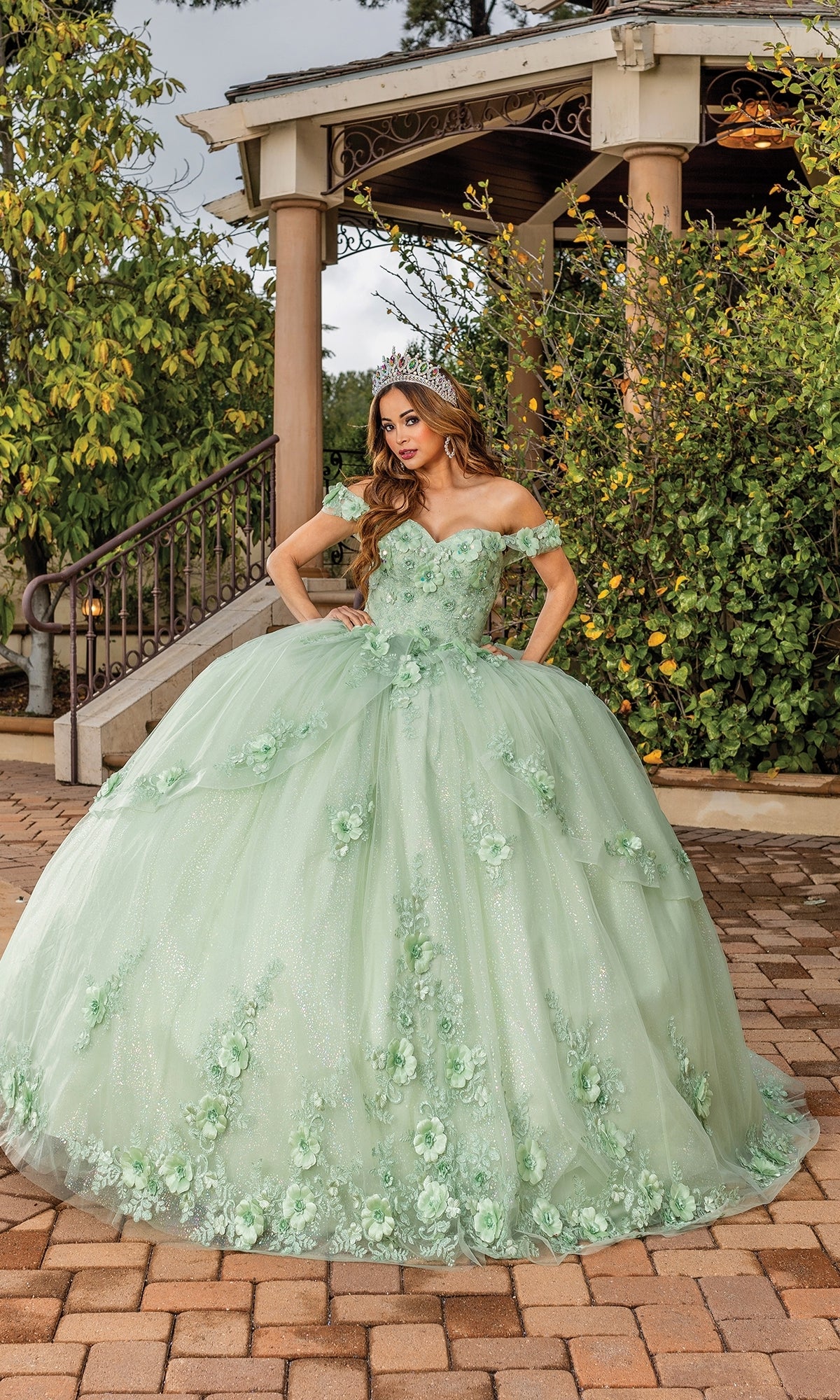 A woman wearing the Quinceanera Dress 1788 By Dancing Queen, a light green A-line ball gown with floral details and off-the-shoulder straps, stands outdoors on a brick path before a gazebo surrounded by greenery.