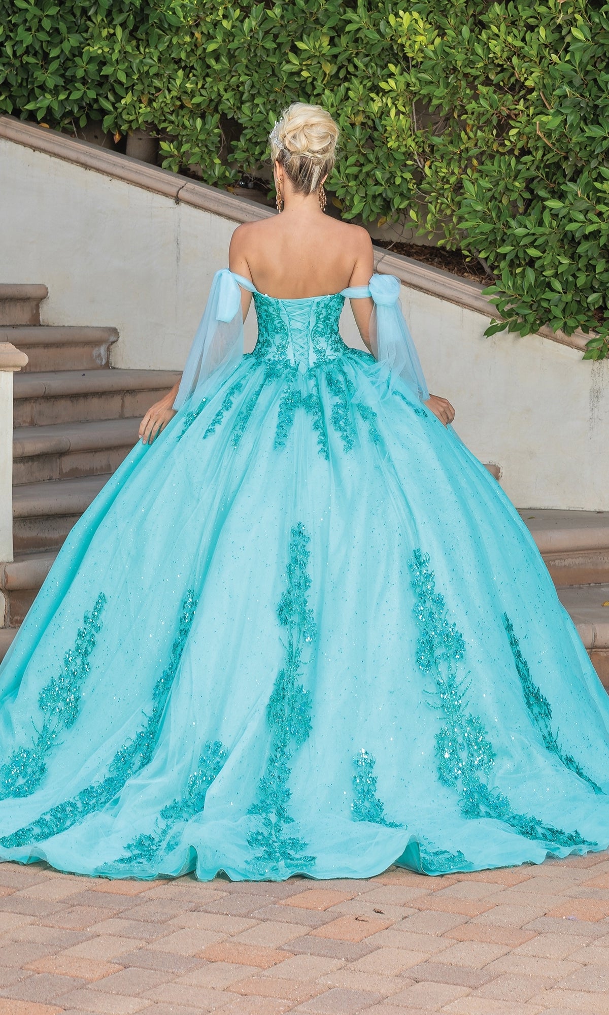 A woman with blonde hair in an updo models the Quinceanera Dress 1791 By Dancing Queen, an off-the-shoulder turquoise ball gown with floral details, standing outside on a tiled walkway near stairs and lush greenery.