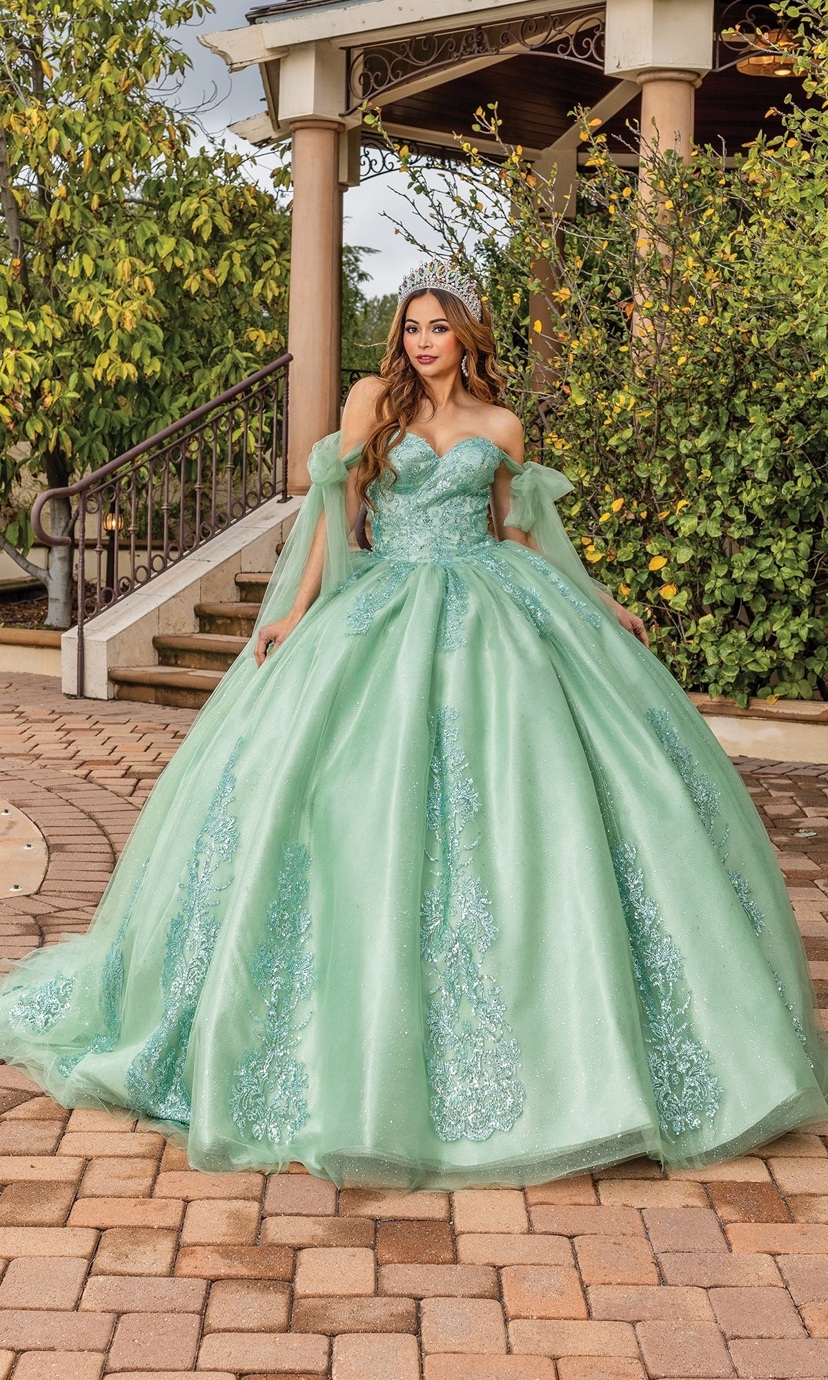 A woman stands outdoors on a stone path in front of greenery and a gazebo, wearing the Quinceanera Dress 1791 By Dancing Queen—a light green, sparkly off-the-shoulder ball gown with off-the-shoulder sleeves.