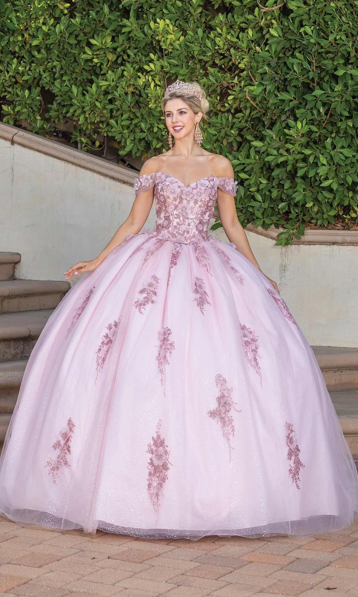 A woman in the Quinceanera Dress 1797 By Dancing Queen, featuring off the shoulder lace applique and floral details, stands on a stone pathway outdoors wearing a tiara, with lush greenery in the background.