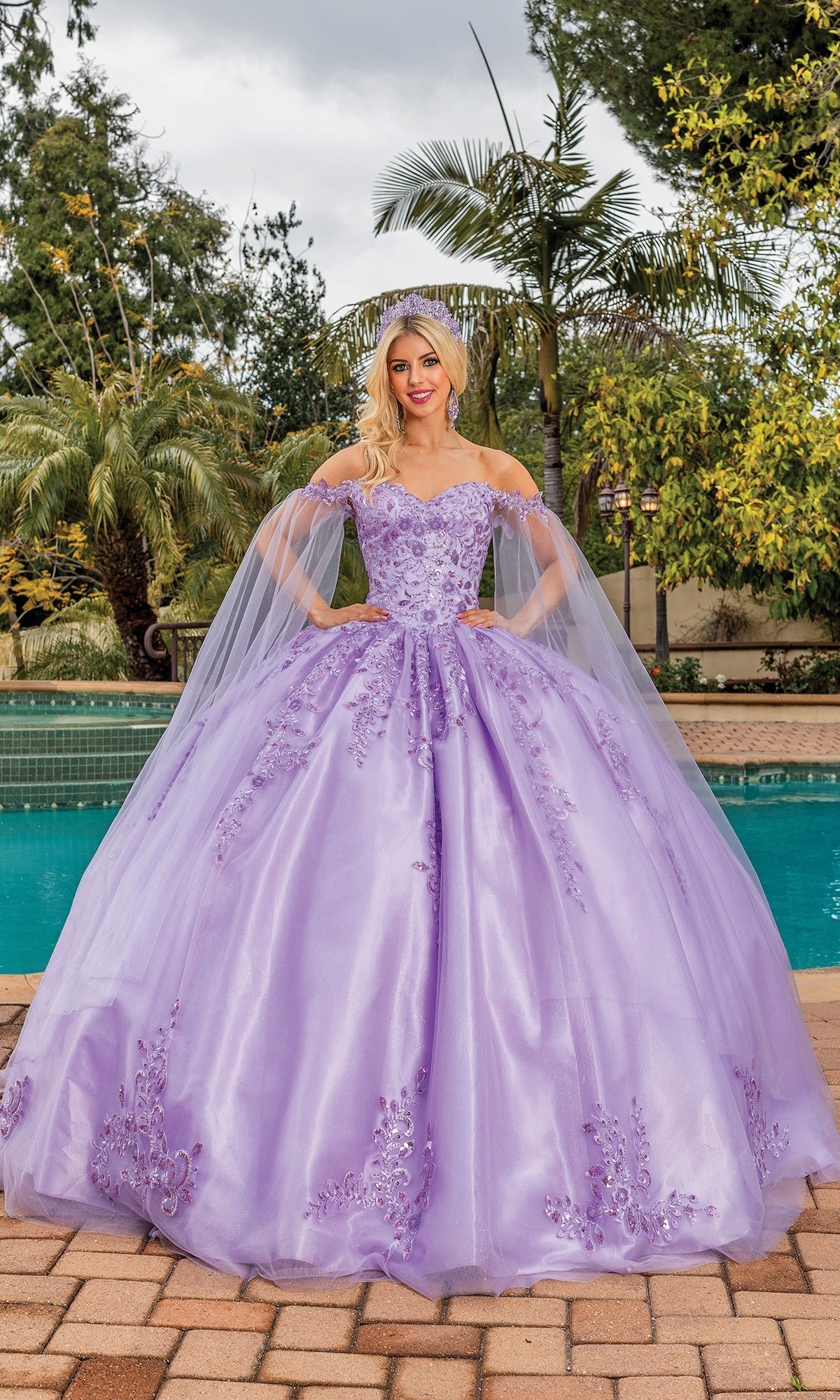 A woman stands outdoors by a pool wearing the Quinceanera Dress 1812 By Dancing Queen, featuring a lavender hue, floral embellishments, off-the-shoulder sleeves, and a full tulle A-line skirt.