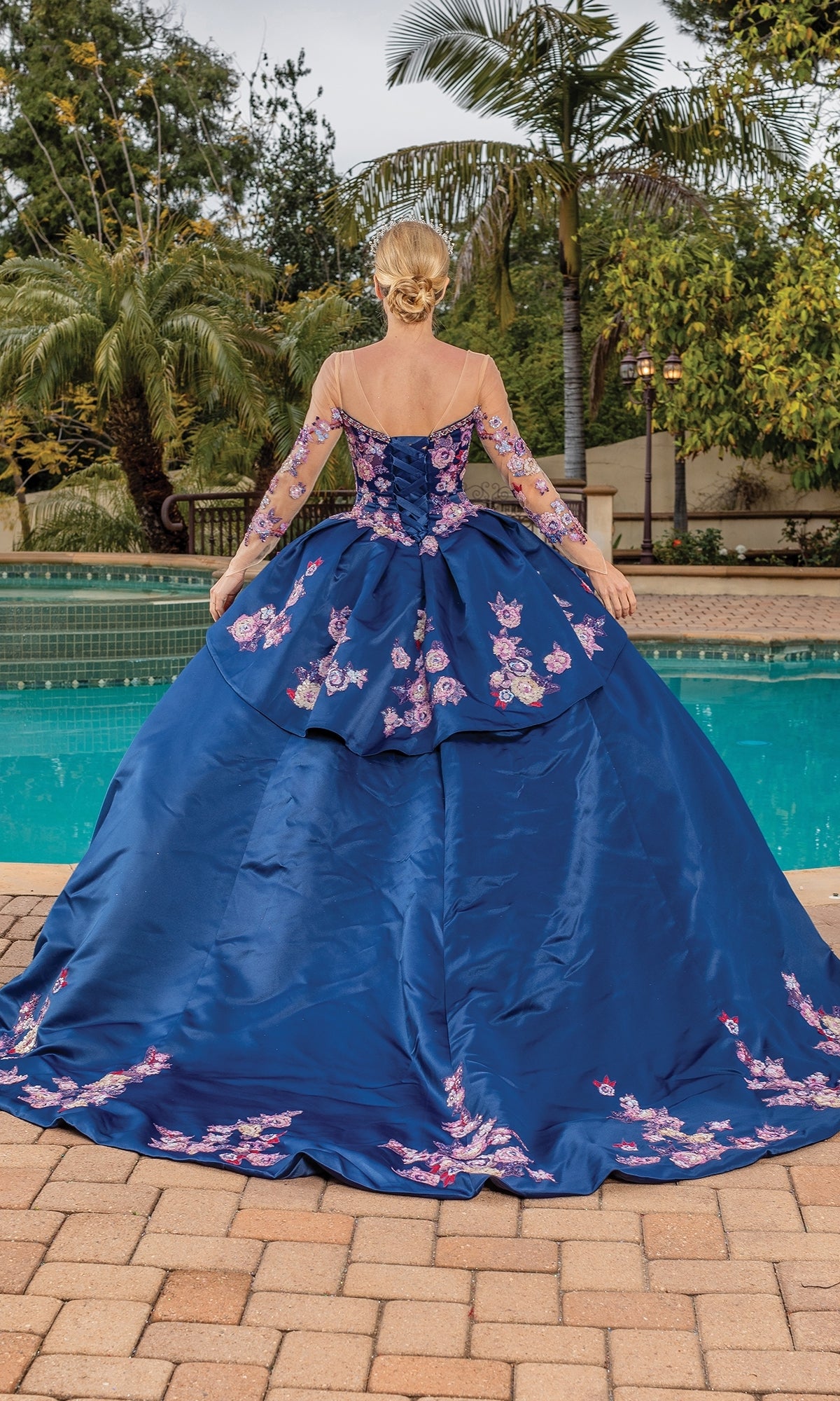 A woman wearing Quinceanera Dress 1816 By Dancing Queen—navy blue with a sweetheart neckline and floral embroidery—stands facing a swimming pool in a lush garden setting.