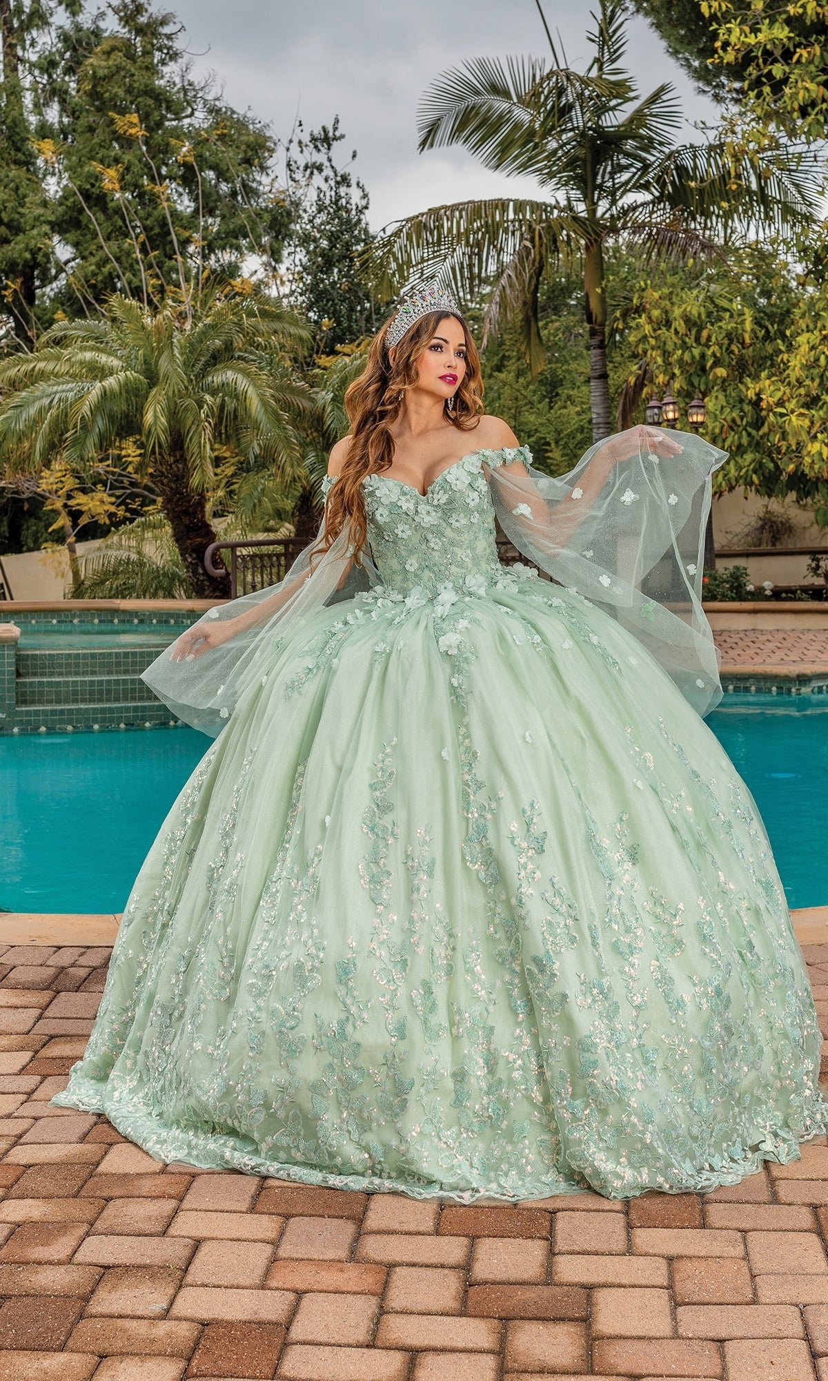 A woman in the Quinceanera Dress 1819 by Dancing Queen, featuring sage green color, long sheer sleeves, and an off-the-shoulder A-line ball gown silhouette, stands on a brick patio near a pool and palm trees.