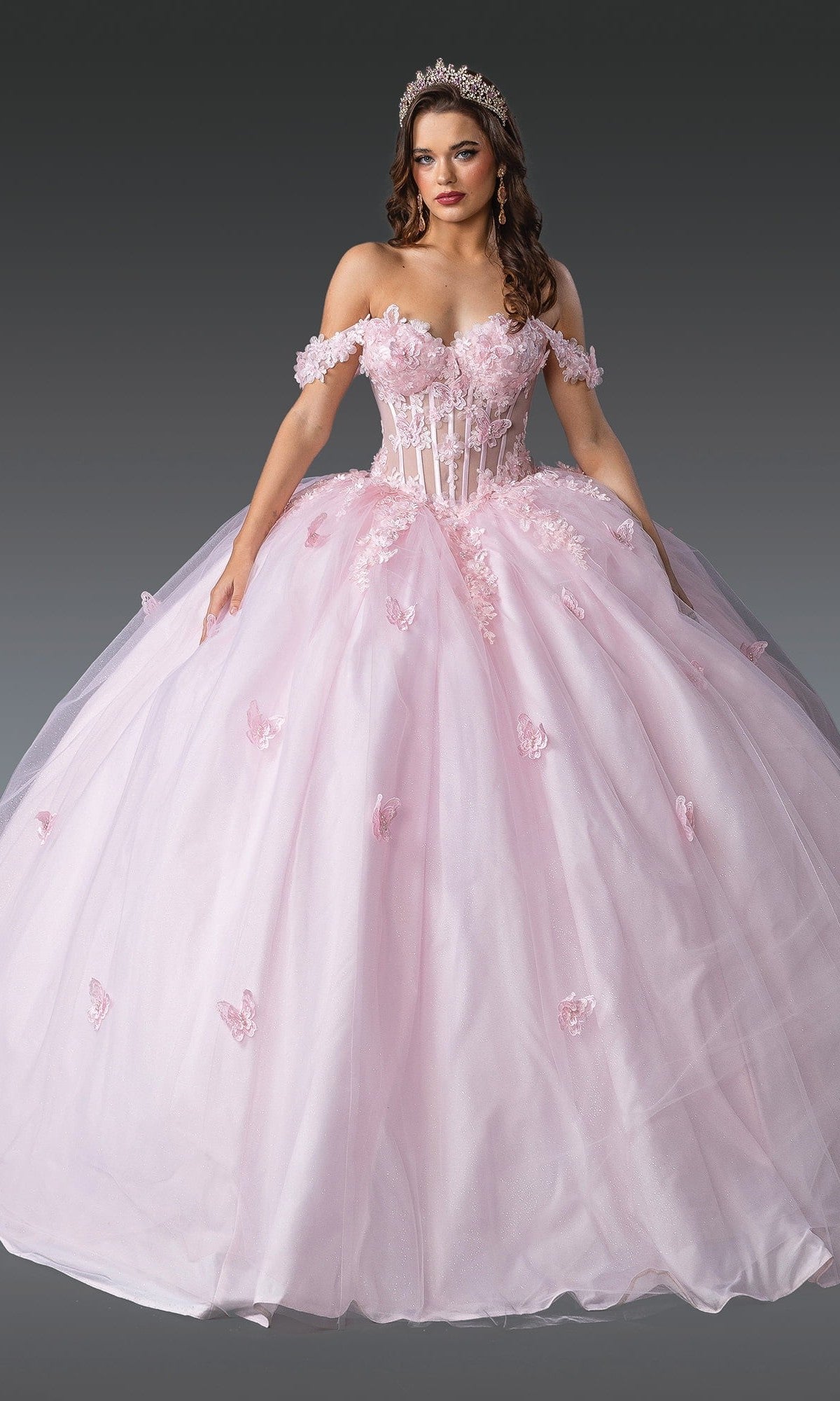 A woman wearing the Quinceanera Dress 1826 By Dancing Queen in blush, featuring off-the-shoulder floral appliqués and a tiara, stands against a dark gray background.