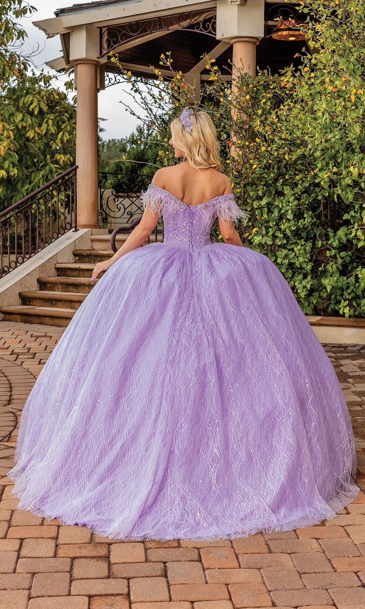 A woman wearing the Quinceanera Dress 1829 By Dancing Queen, an off-the-shoulder lavender ball gown with a sweetheart neckline, stands outdoors with her back to the camera near stone steps and lush greenery.