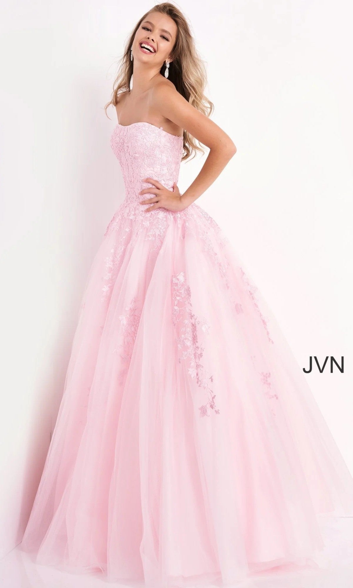 A woman smiles and poses against a plain white background while wearing the Jovani 1831 long formal dress—a strapless, floor-length pastel gown featuring floral detailing. The text “JVN” appears in the corner.