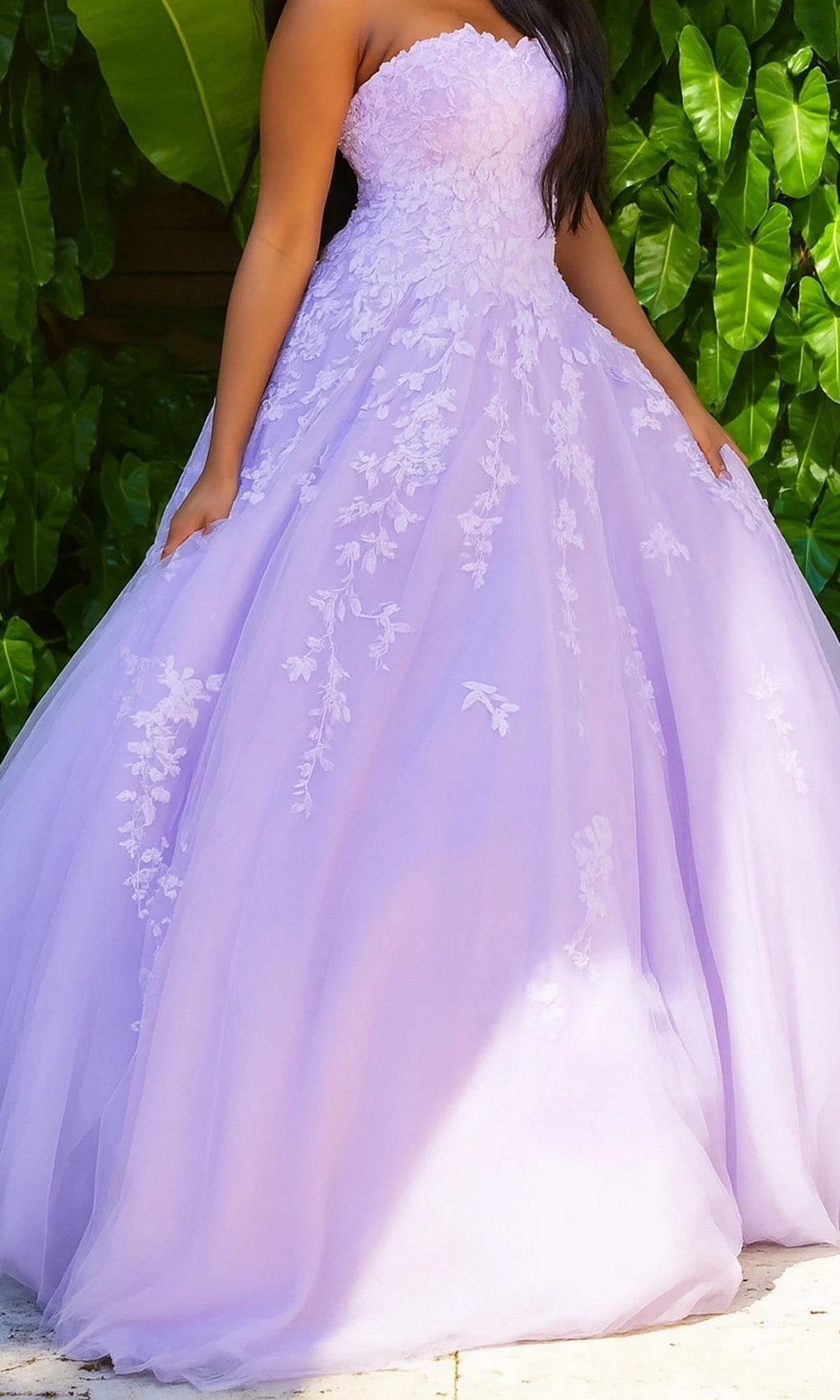 A woman wearing the Jovani 1831 long formal dress in lilac stands in front of large green leaves.