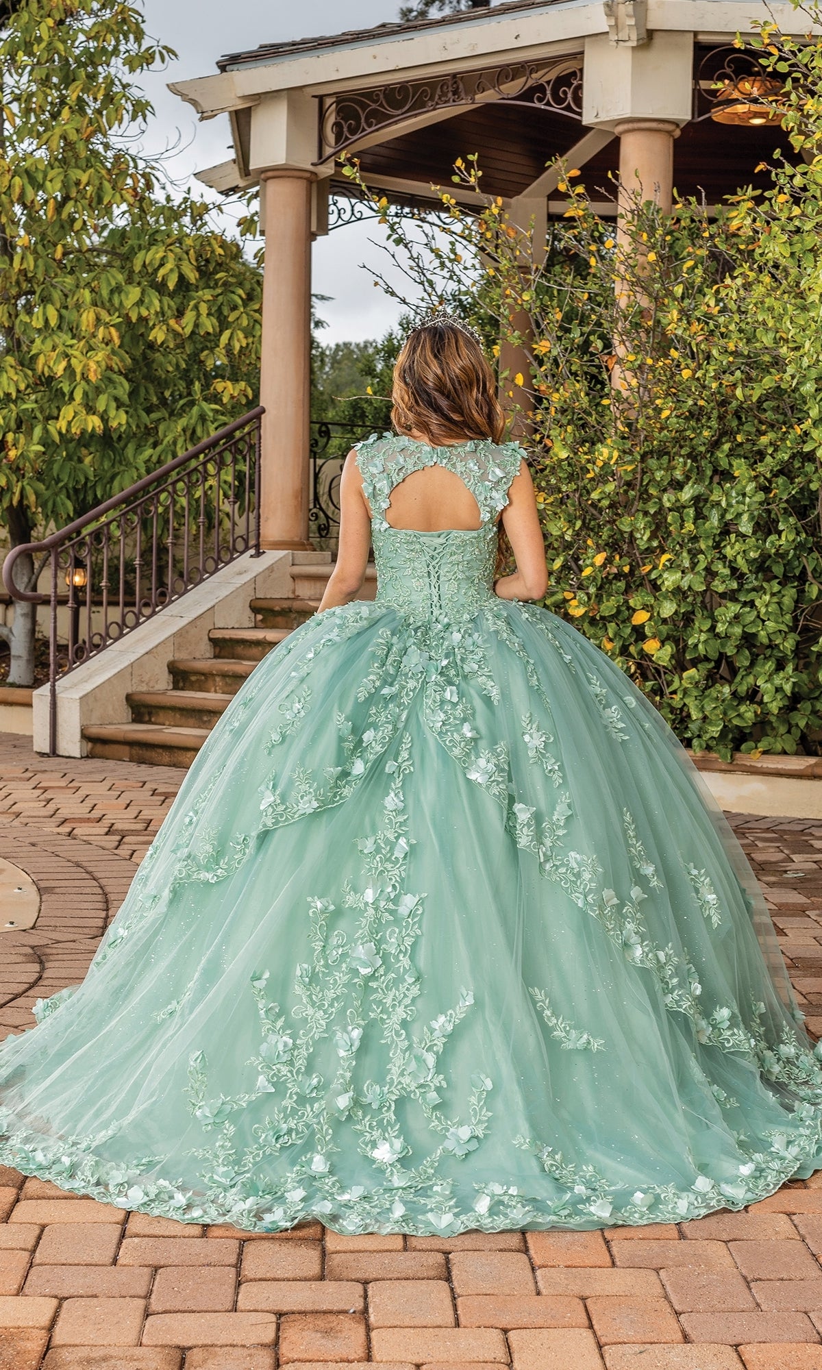 Wearing the Quinceanera Dress 1831 By Dancing Queen in sage green with a sweetheart neckline, a person stands on a brick pathway facing a gazebo, surrounded by lush greenery.