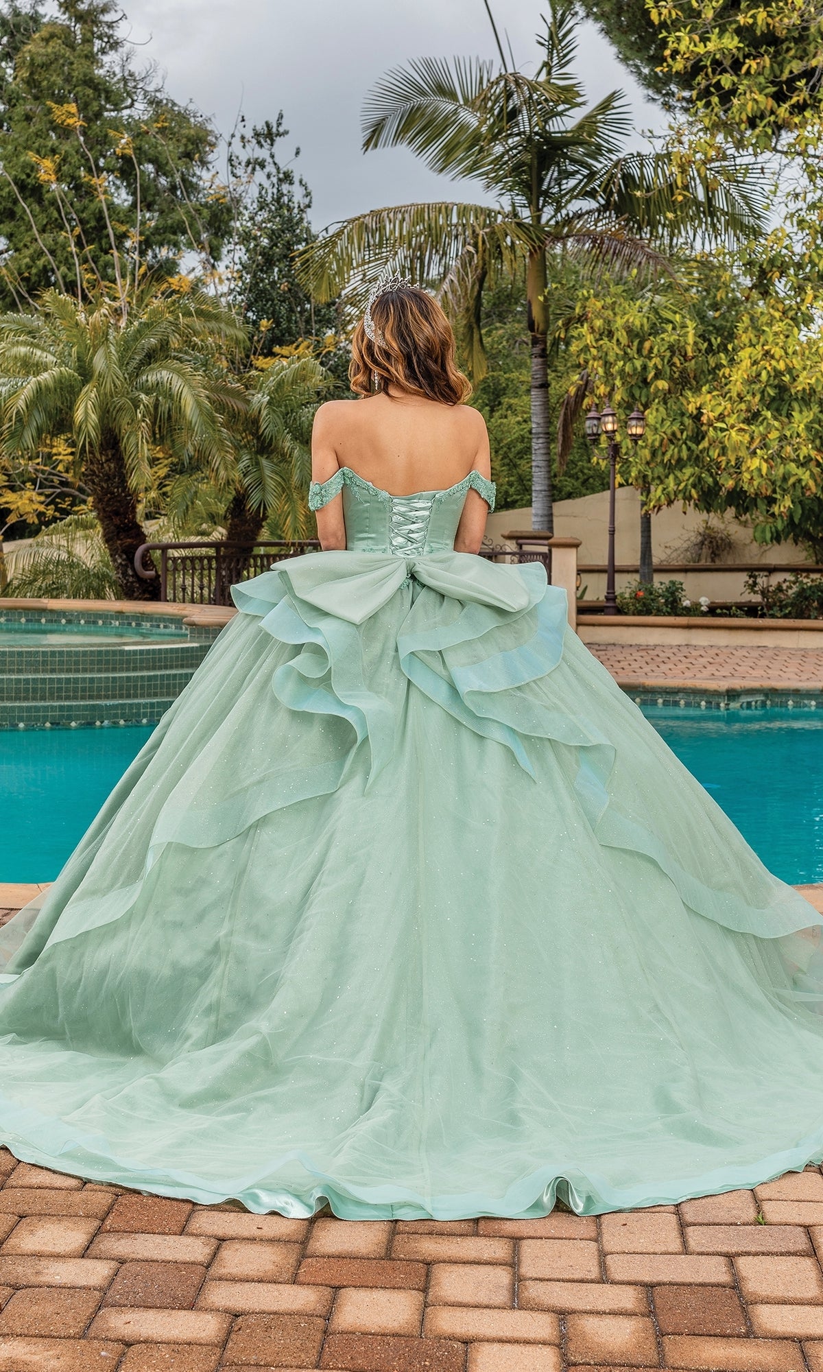 A woman in the Quinceanera Dress 1833 By Dancing Queen, a sage green ball gown with an off-the-shoulder sweetheart neckline, stands by the pool with her back to the camera, surrounded by palm trees and lush greenery.
