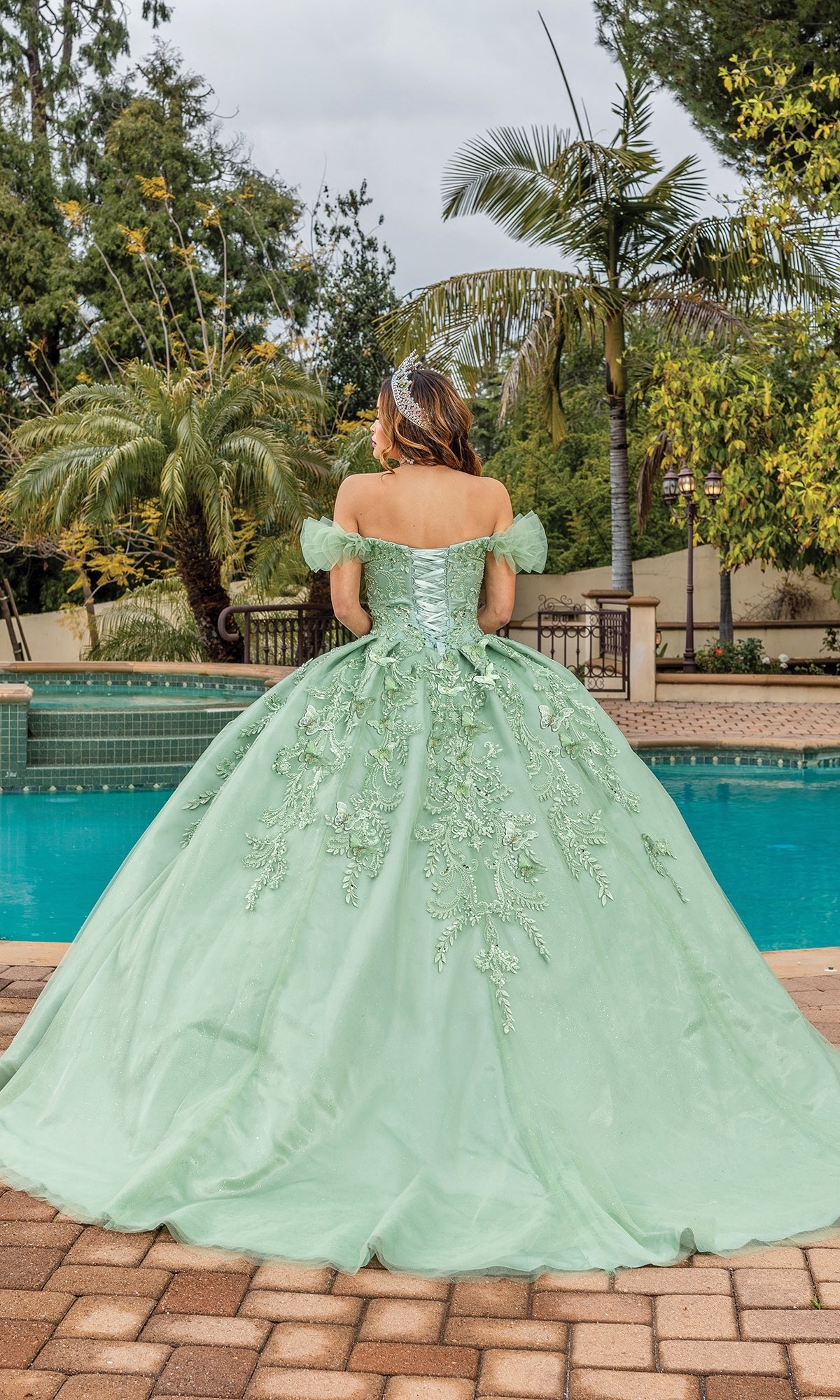 A person in the Quinceanera Dress 1834 By Dancing Queen, a light green off-shoulder gown with a sweetheart neckline, stands by a pool surrounded by palm trees and lush greenery, facing away from the camera.