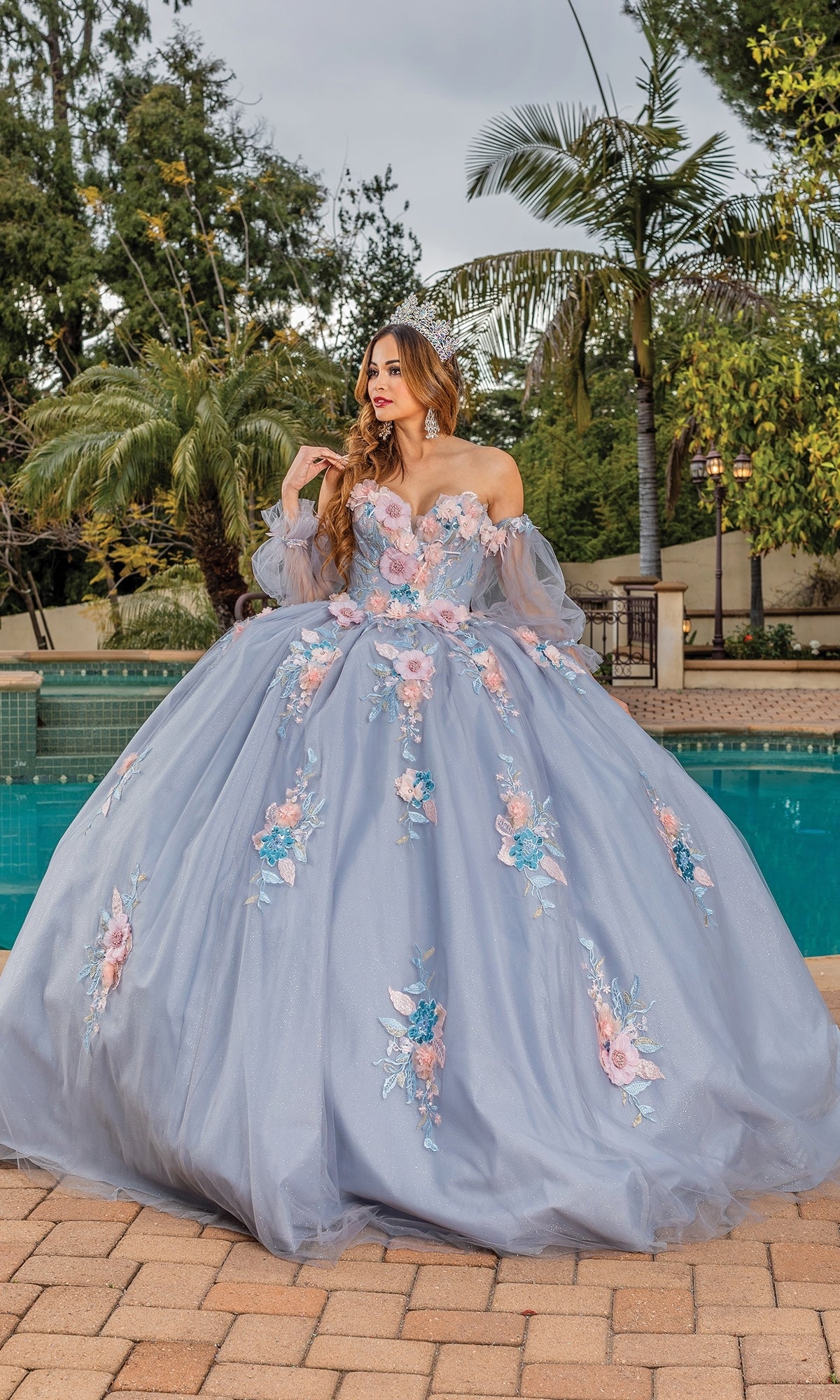 A woman in the Quinceanera Dress 1837 by Dancing Queen, a light blue ball gown with a sweetheart neckline and floral embellishments, stands beside a pool among palm trees and lush greenery.