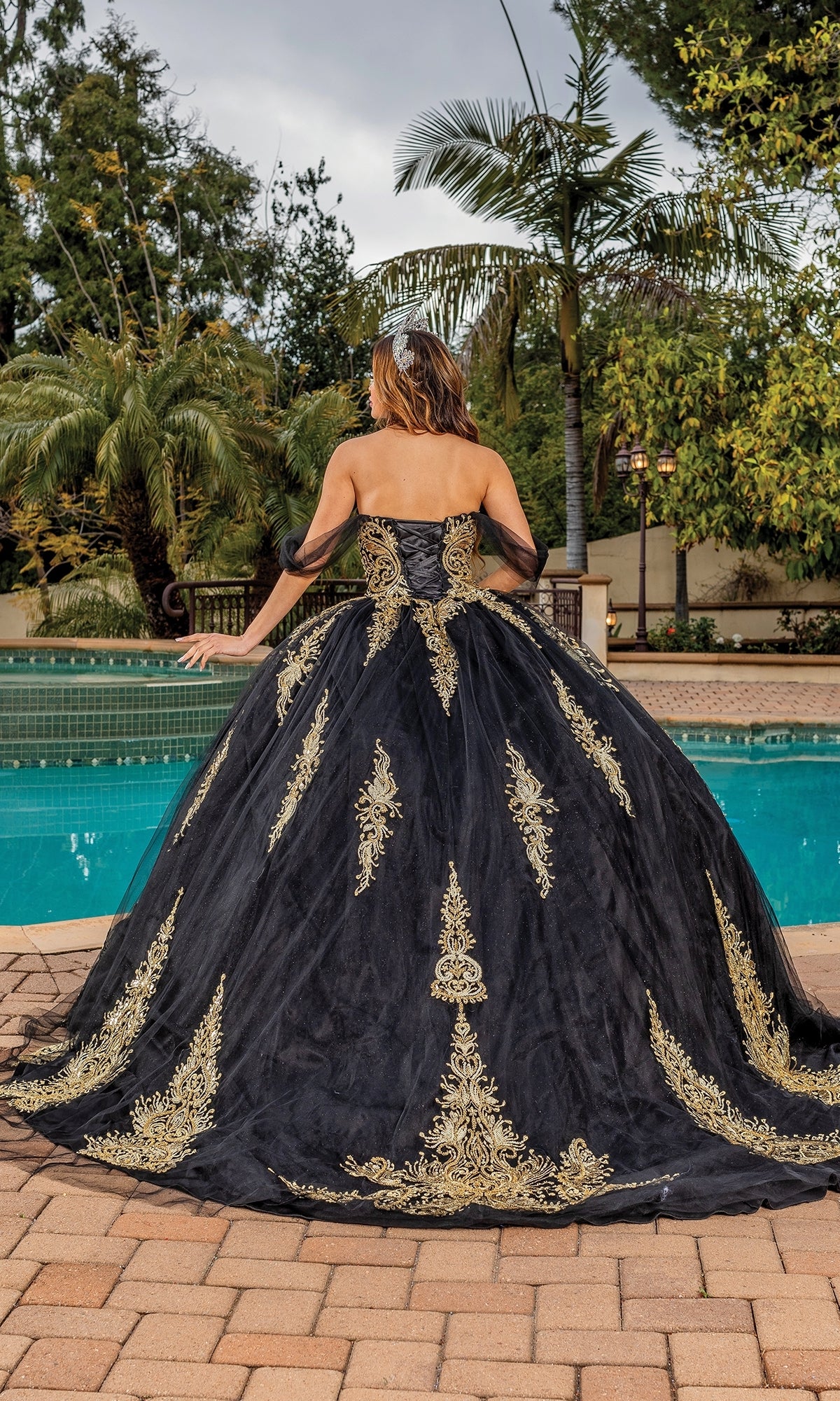 A woman wearing the Quinceanera Dress 1839 By Dancing Queen, with gold lace applique and an off the shoulder design, stands by a pool with her back to the camera, surrounded by lush trees and greenery.