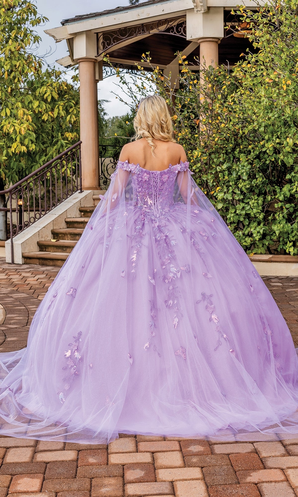A person with blonde hair stands outdoors, facing away from the camera, wearing the Quinceanera Dress 1840 By Dancing Queen—a long, lavender off-the-shoulder ball gown with floral details.
