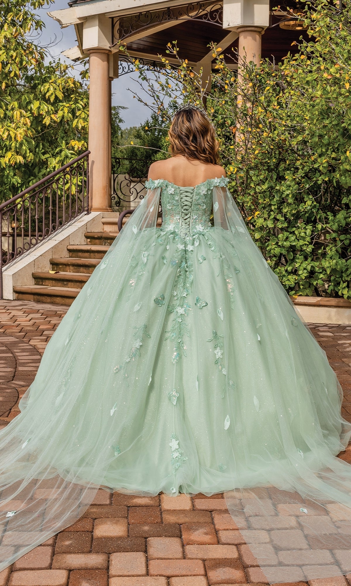 A woman stands outdoors near a staircase and lush greenery, wearing the Quinceanera Dress 1840 By Dancing Queen—a light green, off-the-shoulder gown with floral details, a sweetheart neckline, and a long train.