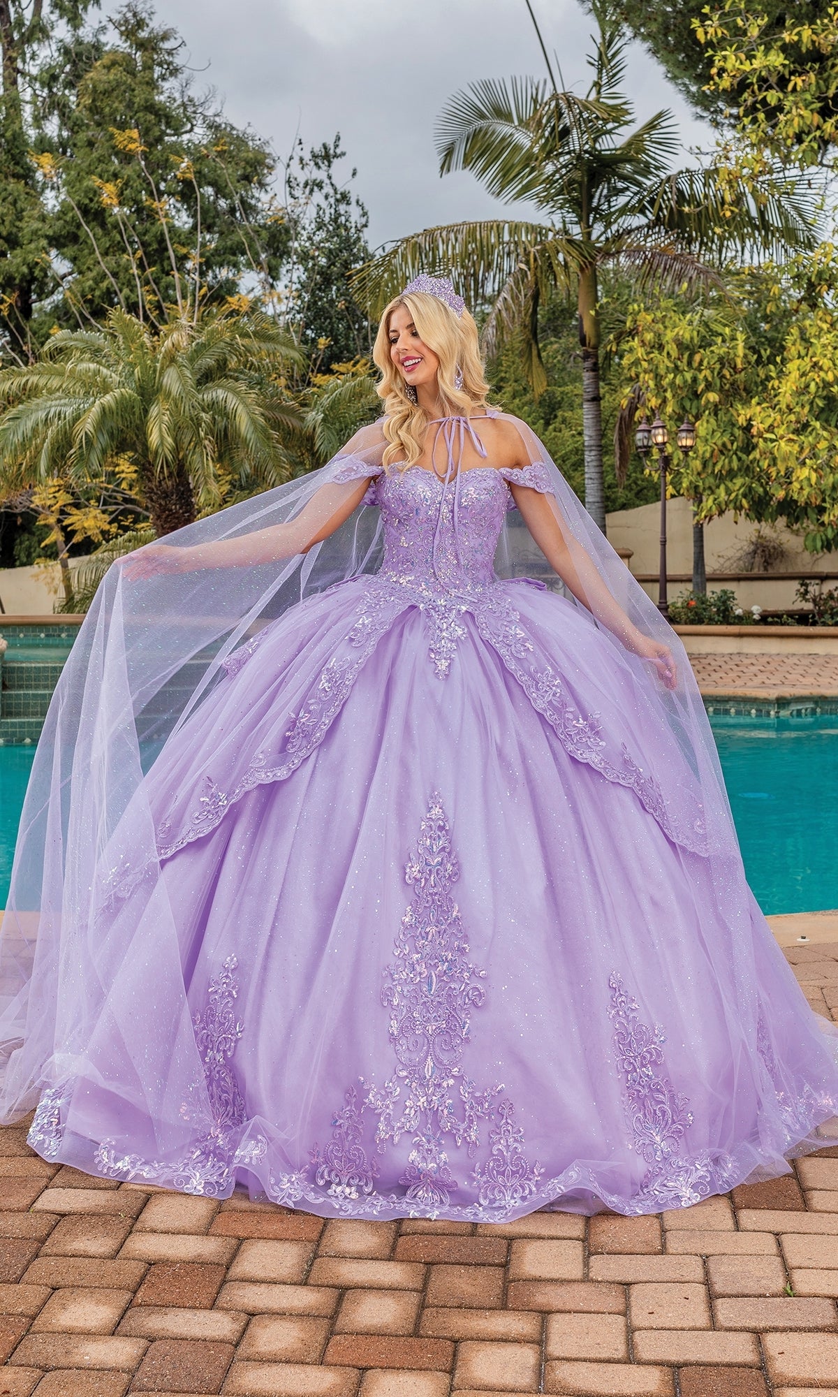 A woman stands by a pool, wearing the Quinceanera Dress 1843 By Dancing Queen—an elegant lavender A-line gown with intricate embroidery, sheer removable cape sleeves, and off-the-shoulder straps. Palm trees and greenery are in the background.