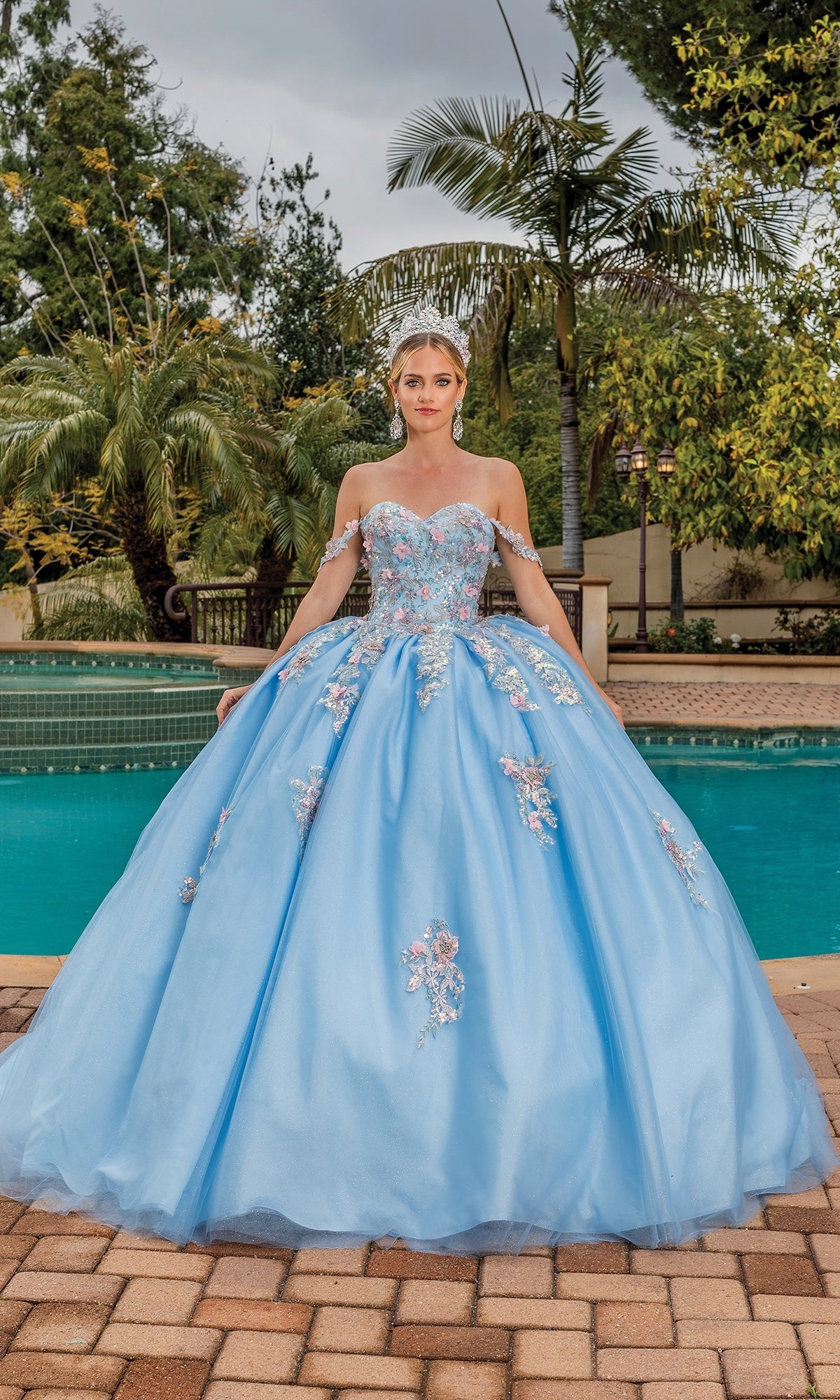 A woman stands by a pool in the Quinceanera Dress 1845 By Dancing Queen, a light blue ball gown with floral embellishments, surrounded by lush tropical trees and plants.