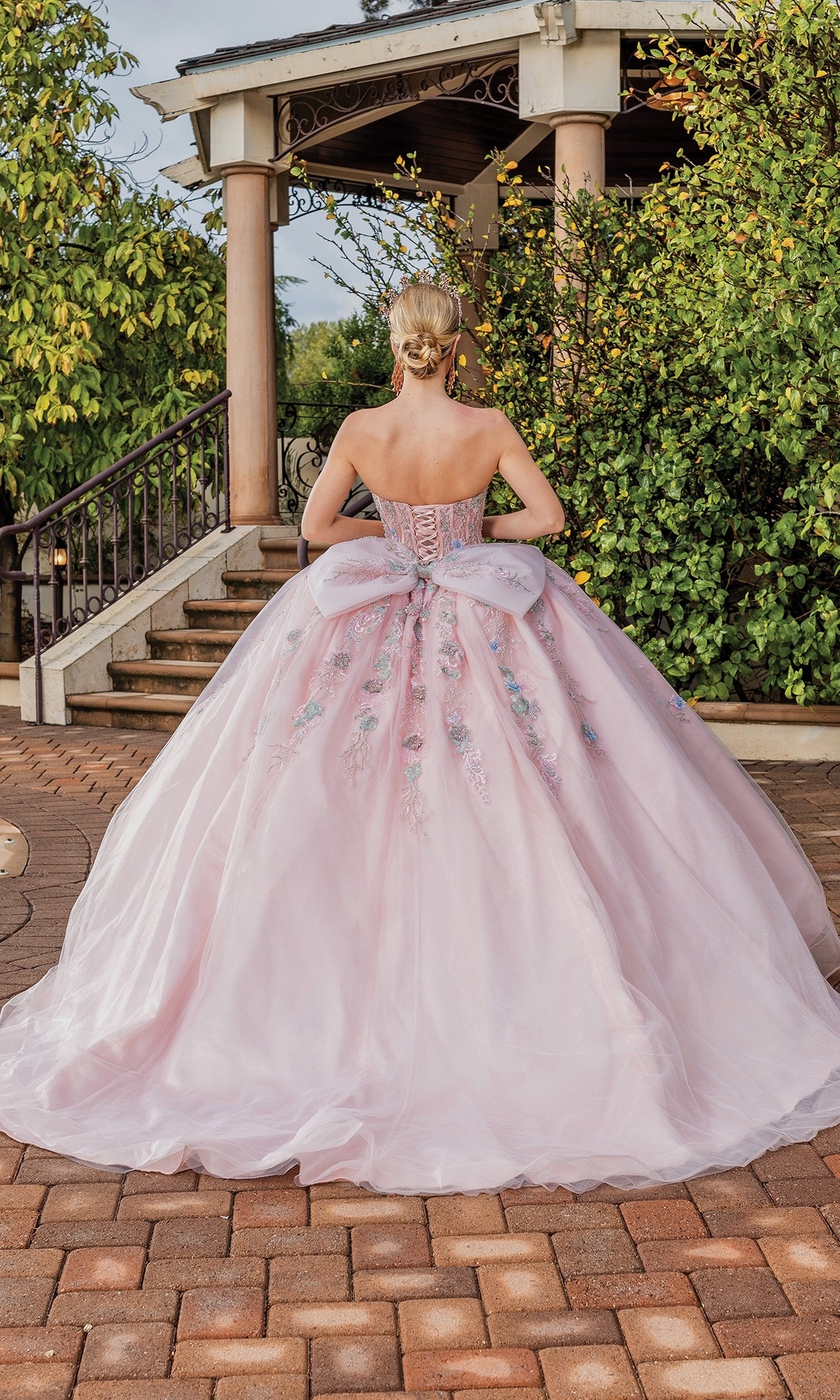 A woman faces away in front of a garden gazebo wearing the Quinceanera Dress 1848 By Dancing Queen, a light pink A-line ball gown with floral embroidery and a dramatic bow back.