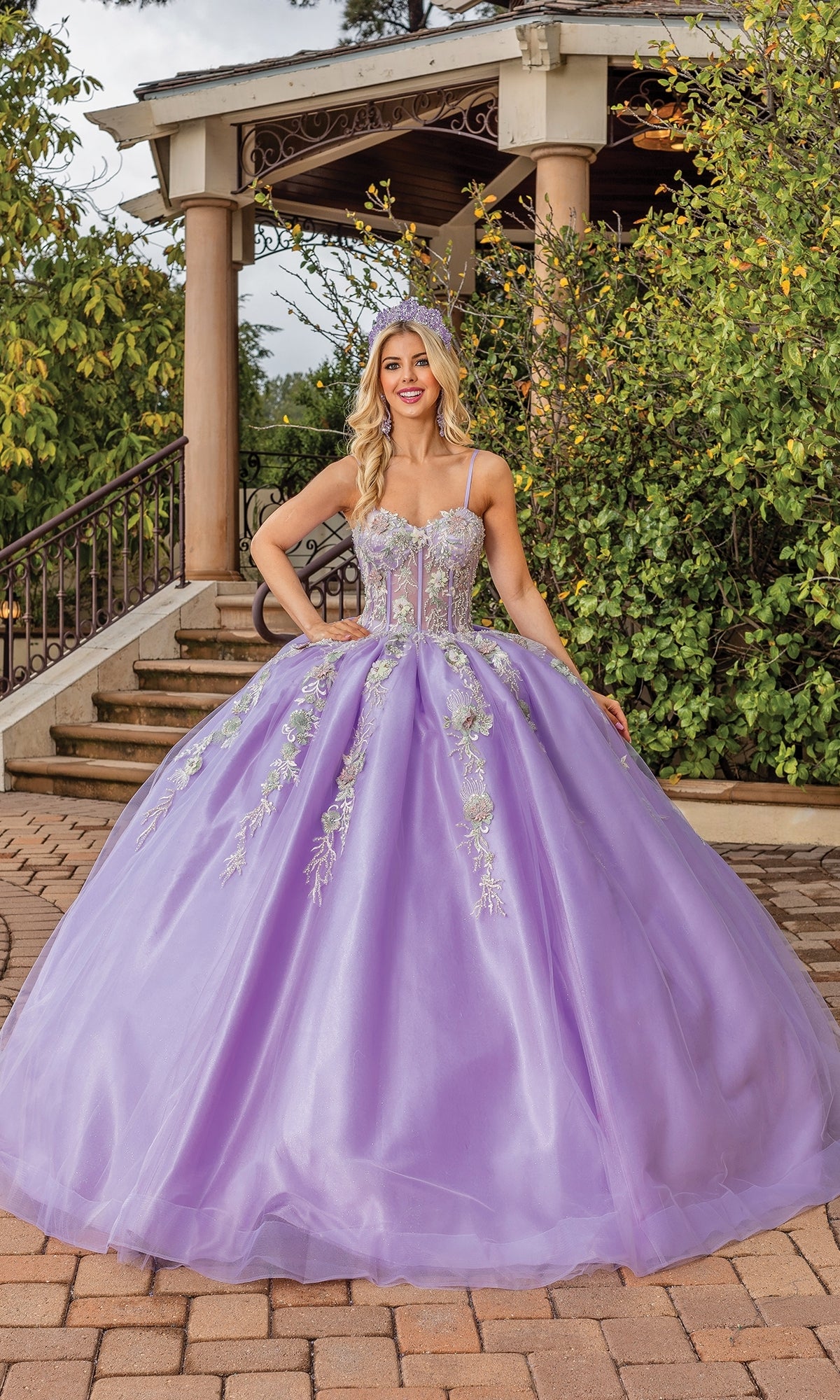 A woman poses outdoors in front of a gazebo and greenery, wearing the Quinceanera Dress 1848 By Dancing Queen—a lavender a-line ball gown with floral embroidery and a sweetheart neckline.