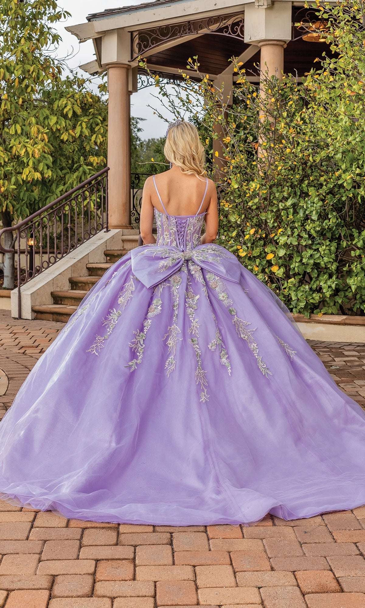 A woman stands outdoors in the Quinceanera Dress 1848 By Dancing Queen, an a-line lavender ball gown with floral embroidery and a large bow back, her elegant silhouette captured from behind.