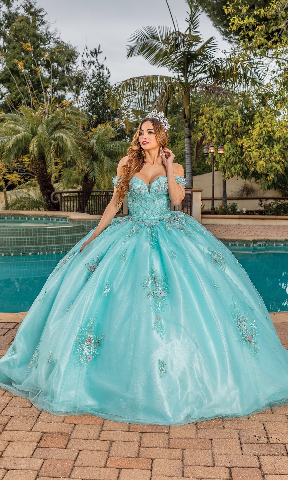 A woman wears the Quinceanera Dress 1850 By Dancing Queen—an off-the-shoulder aqua blue ball gown with floral embroidery—while standing on a brick patio by a pool, surrounded by palm trees and lush greenery.