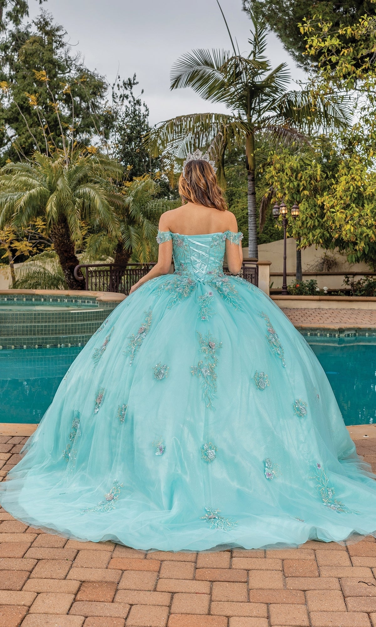 A young woman stands by a pool in the Quinceanera Dress 1850 By Dancing Queen, featuring an aqua blue ball gown with floral embellishments and an off-the-shoulder, sweetheart neckline, her back to the camera.
