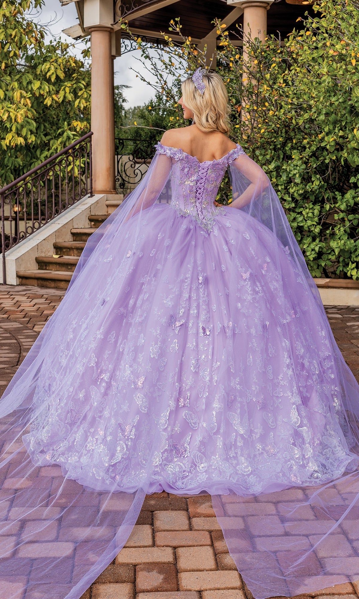 A woman stands outdoors on a stone path, facing away, wearing the Quinceanera Dress 1852 By Dancing Queen—an off-the-shoulder A-line ball gown with a full skirt and long draped cape sleeves.