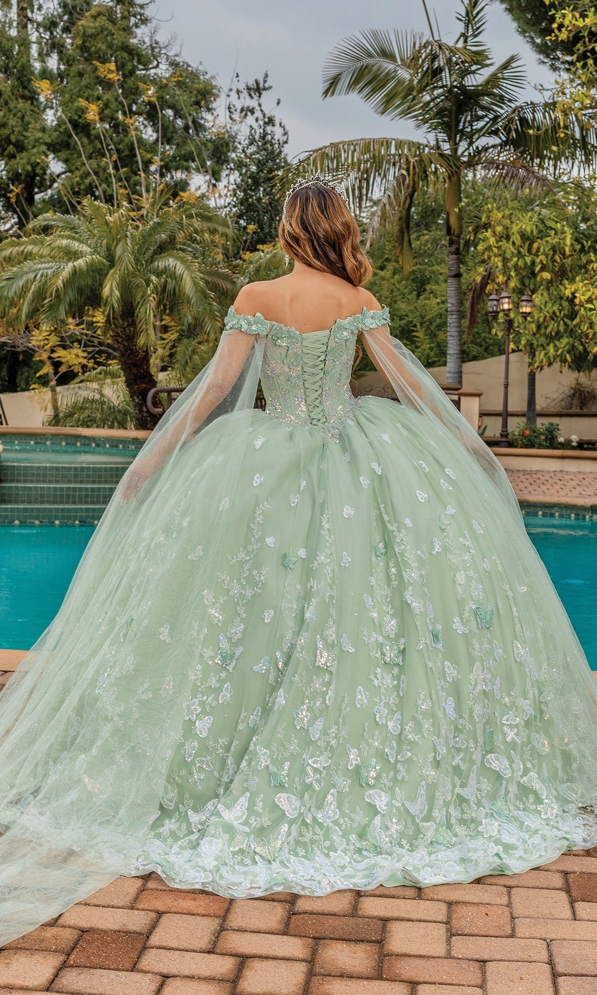 A woman wearing the Quinceanera Dress 1852 By Dancing Queen, featuring an a-line silhouette, draped cape sleeves, and floral details, stands by a pool with her back to the camera, surrounded by palm trees.