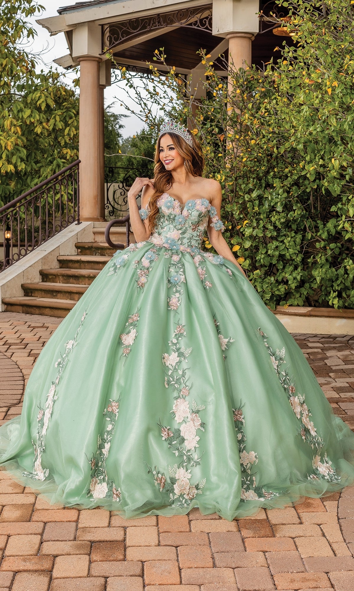 A woman stands outdoors in the Quinceanera Dress 1856 By Dancing Queen, a sage green gown with floral embellishments and a tiara, posing in front of steps and greenery.