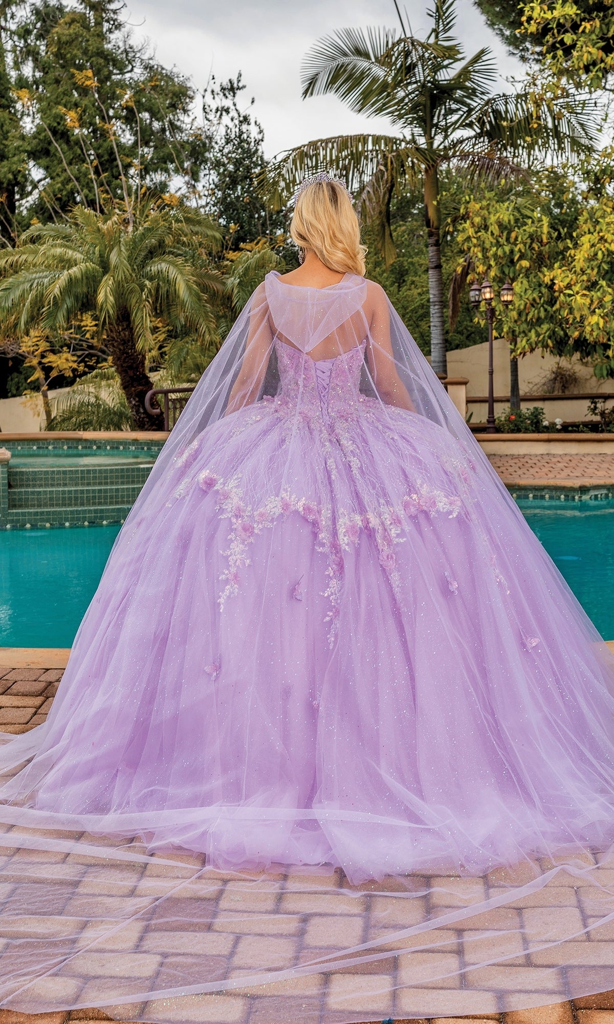 A person in the Quinceanera Dress 1858 By Dancing Queen, featuring a lilac hue, sweetheart neckline, and tulle overlay, stands by a pool with their back to the camera amid lush greenery and palm trees.
