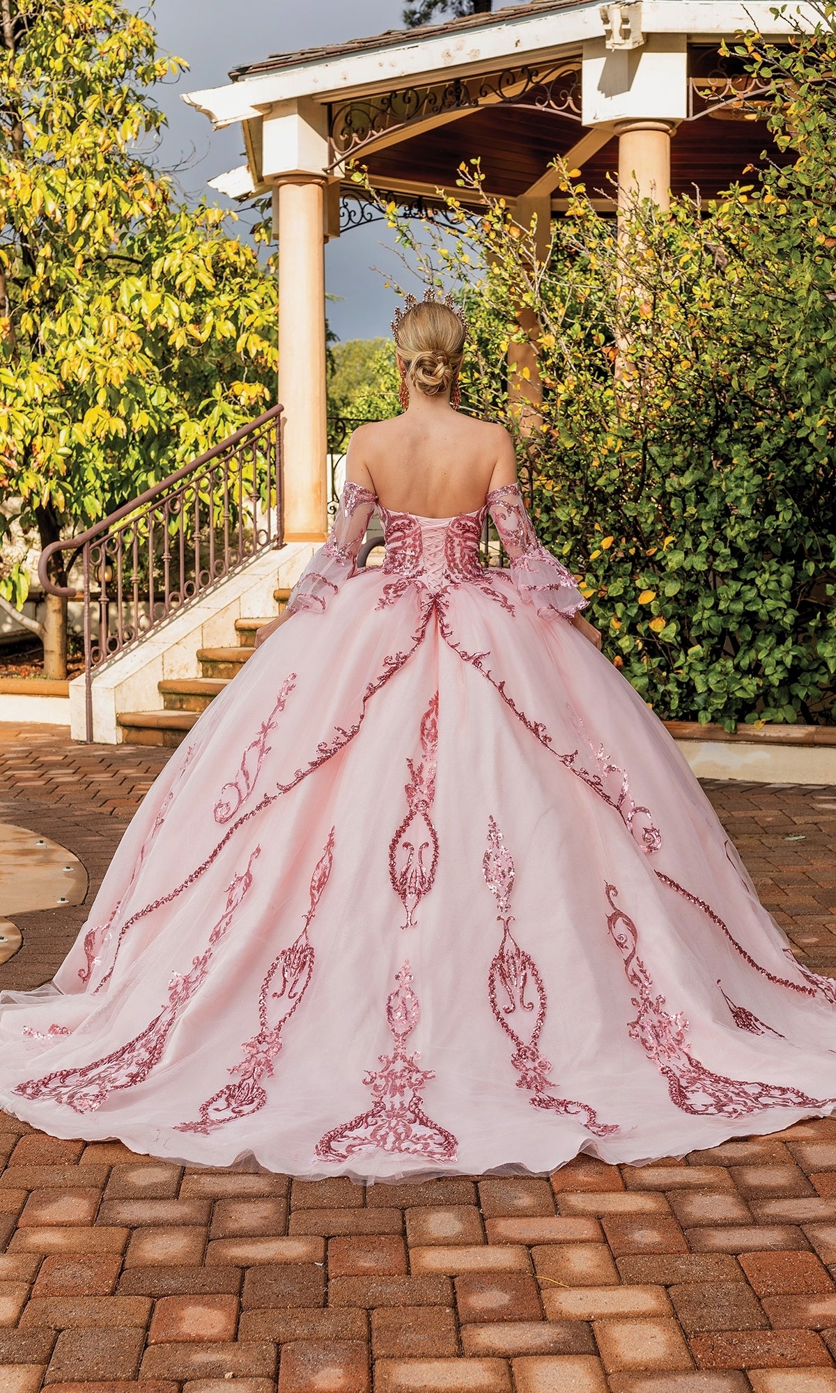 A woman wearing the Quinceanera Dress 1861 By Dancing Queen in blush pink with red embroidery stands on a brick path, facing away toward a gazebo amid greenery.
