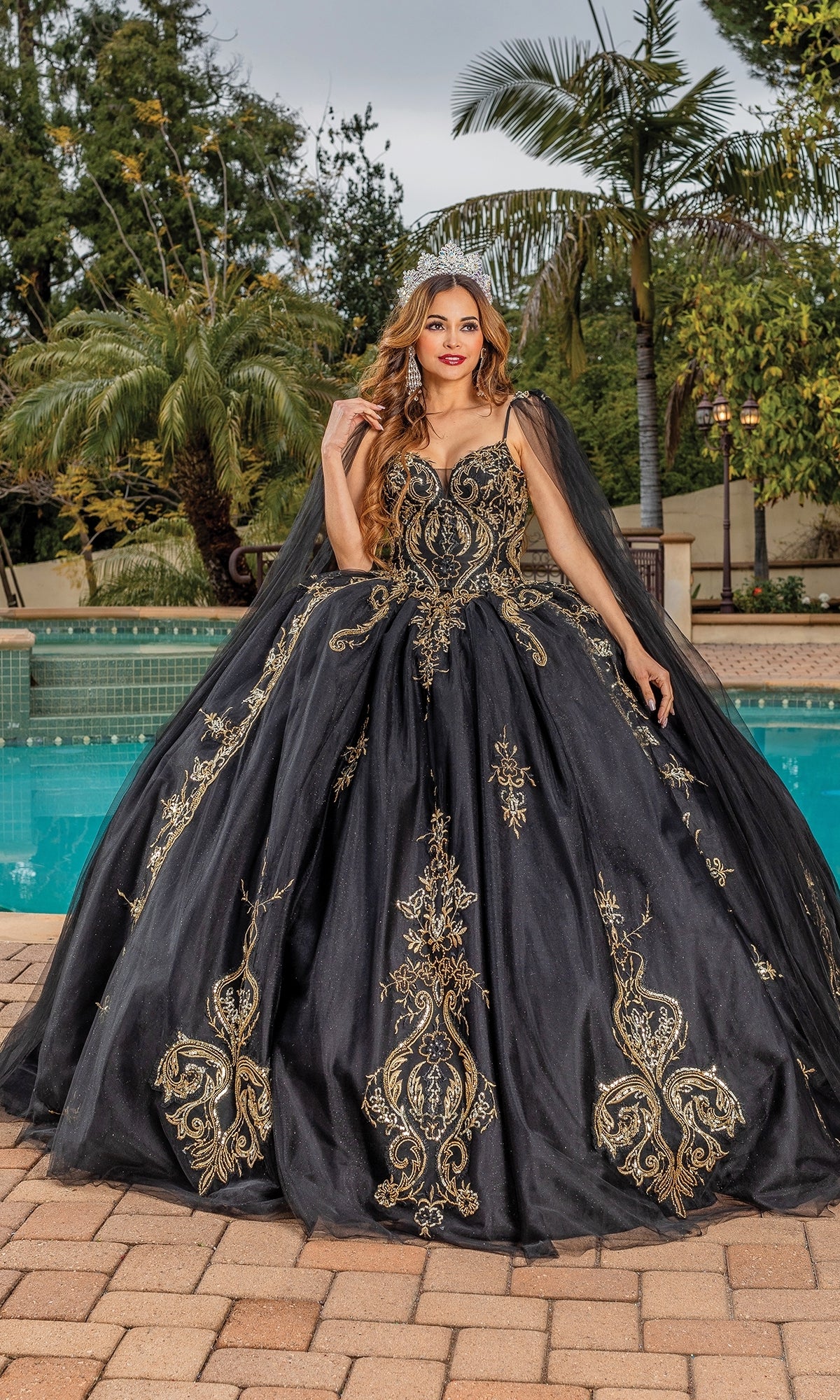 A woman wearing the Quinceanera Dress 1866 By Dancing Queen, featuring black fabric with gold embroidery and a sweetheart neckline, stands by a swimming pool amid palm trees and lush greenery.