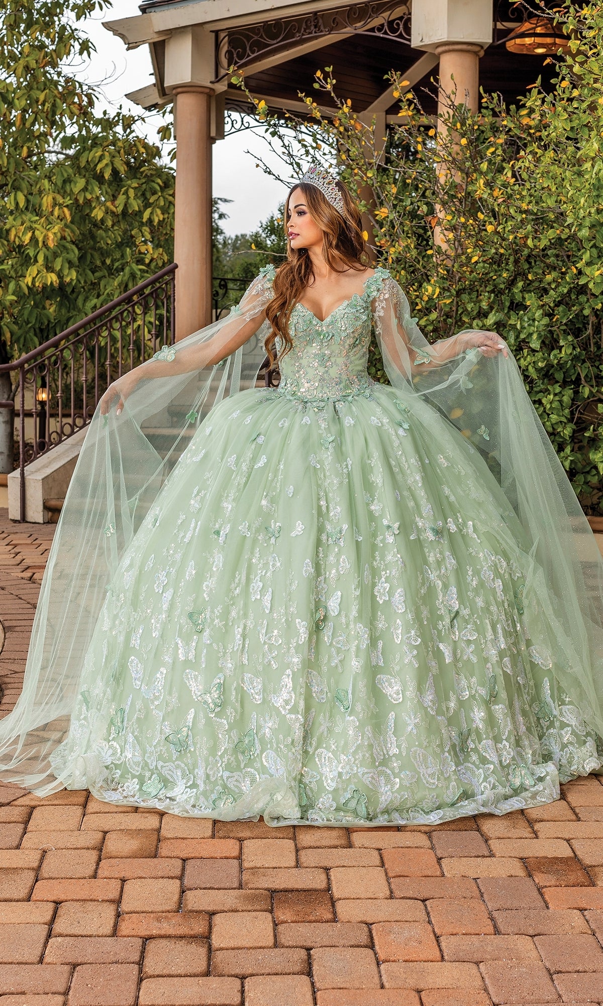 A woman stands outdoors on a brick path, holding out her Quinceanera Dress 1878 By Dancing Queen—a light green, embellished ball gown with sheer sleeves and a sweetheart neckline.