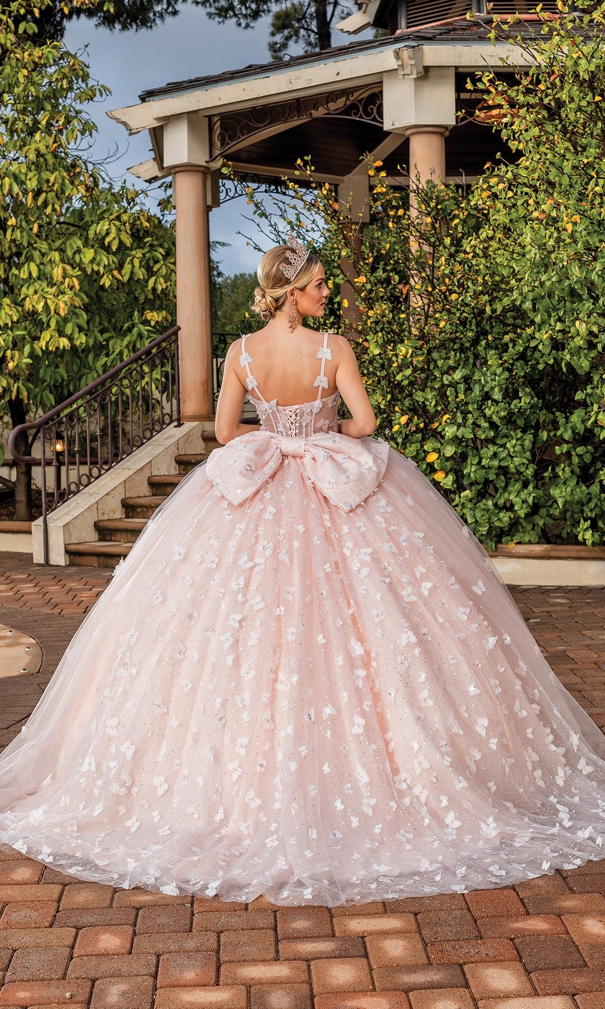 A young woman stands outdoors before a gazebo, her back to the camera, wearing the Quinceanera Dress 1879 By Dancing Queen—a pink ball gown with a large bow and floral appliqués.
