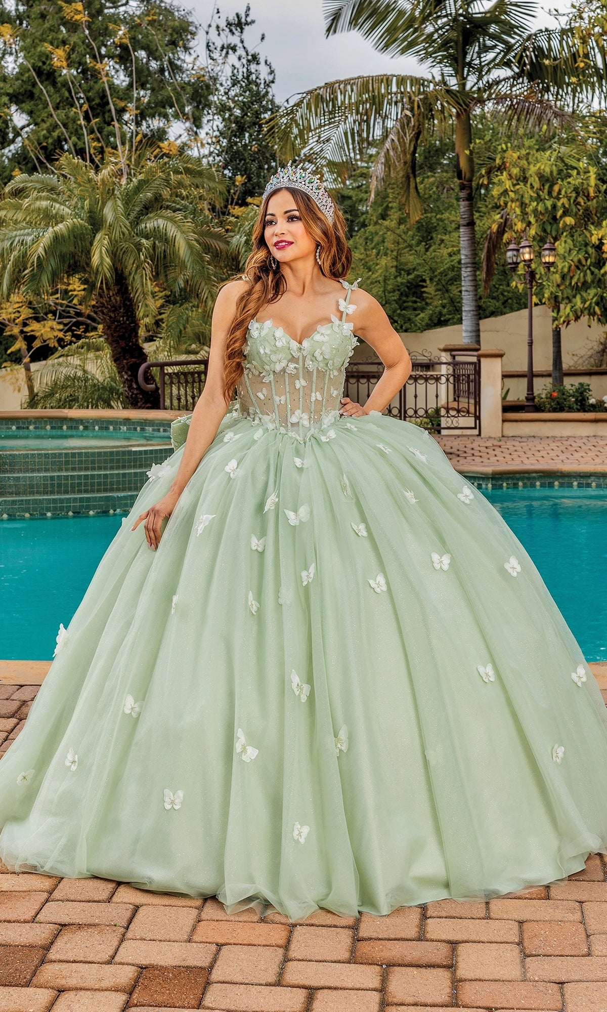 A woman wearing the Quinceanera Dress 1879 by Dancing Queen—a light green ball gown with floral details and a tiara—smiles as she stands by a swimming pool.