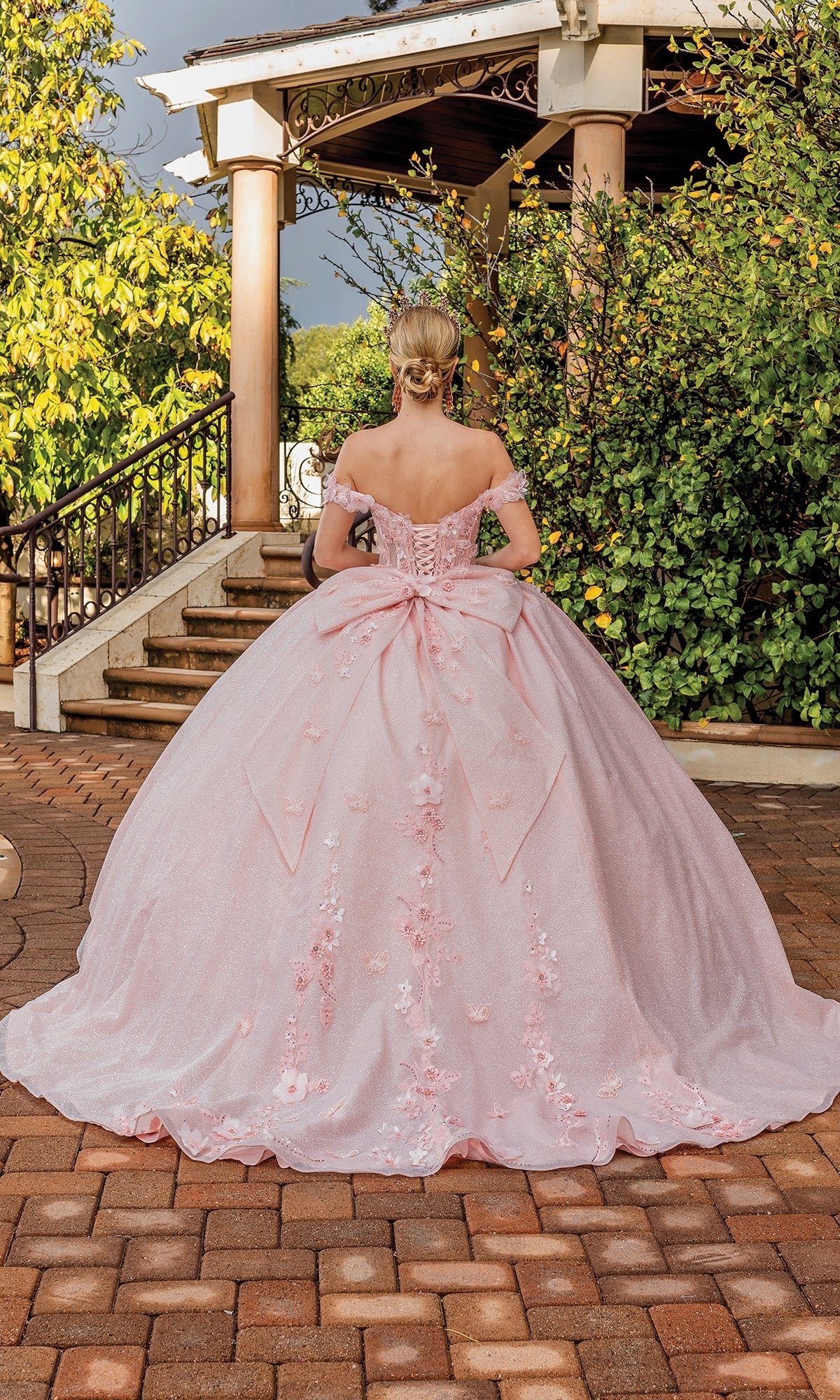 A woman in the Quinceanera Dress 1888 By Dancing Queen—a pink off-the-shoulder A-line ball gown with floral details and a large bow—stands outdoors facing a staircase surrounded by lush greenery.