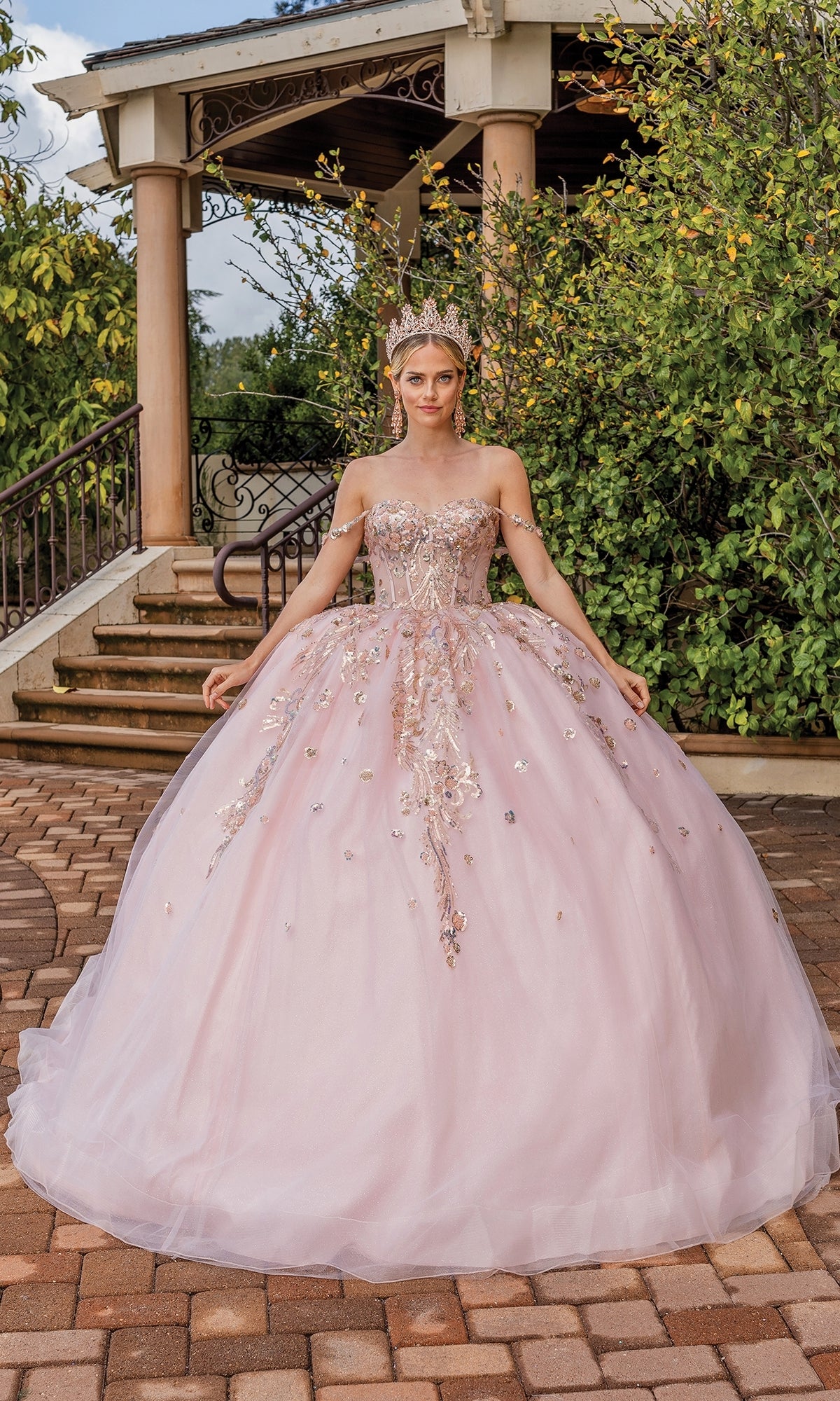 A woman poses outdoors in front of stone steps and greenery, wearing the Quinceanera Dress 1894 By Dancing Queen—an a-line pink ball gown with floral accents, jeweled crown, and sweetheart neckline.