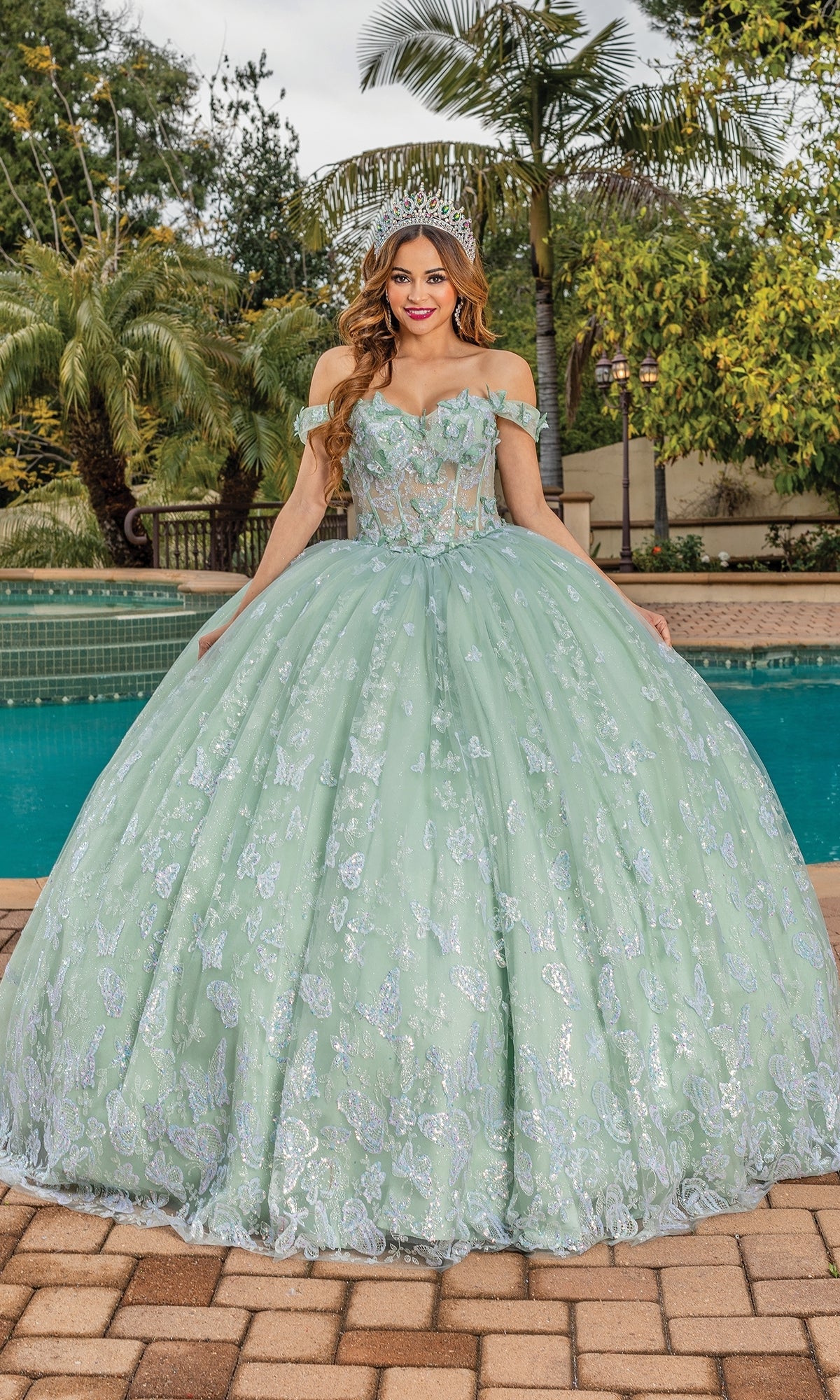 A woman stands by a pool in front of palm trees, wearing the Quinceanera Dress 1897 By Dancing Queen—a pastel off-shoulder gown with silver embroidery and a sparkling silver tiara.