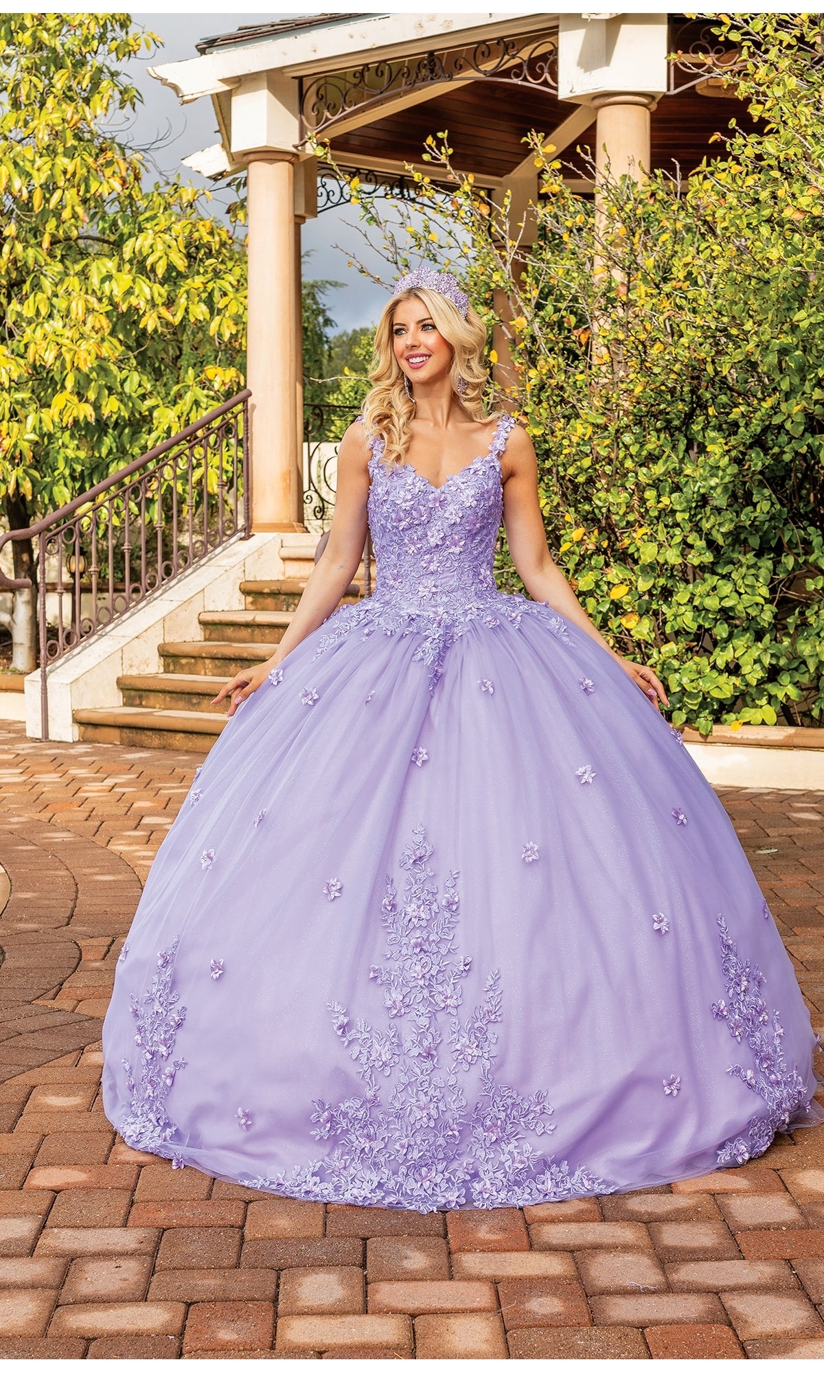 A woman stands outdoors in front of a gazebo and greenery, holding the sides of her skirt while wearing the Quinceanera Dress 1898 By Dancing Queen, a lavender A-line ball gown with floral embellishments and a V-neckline.