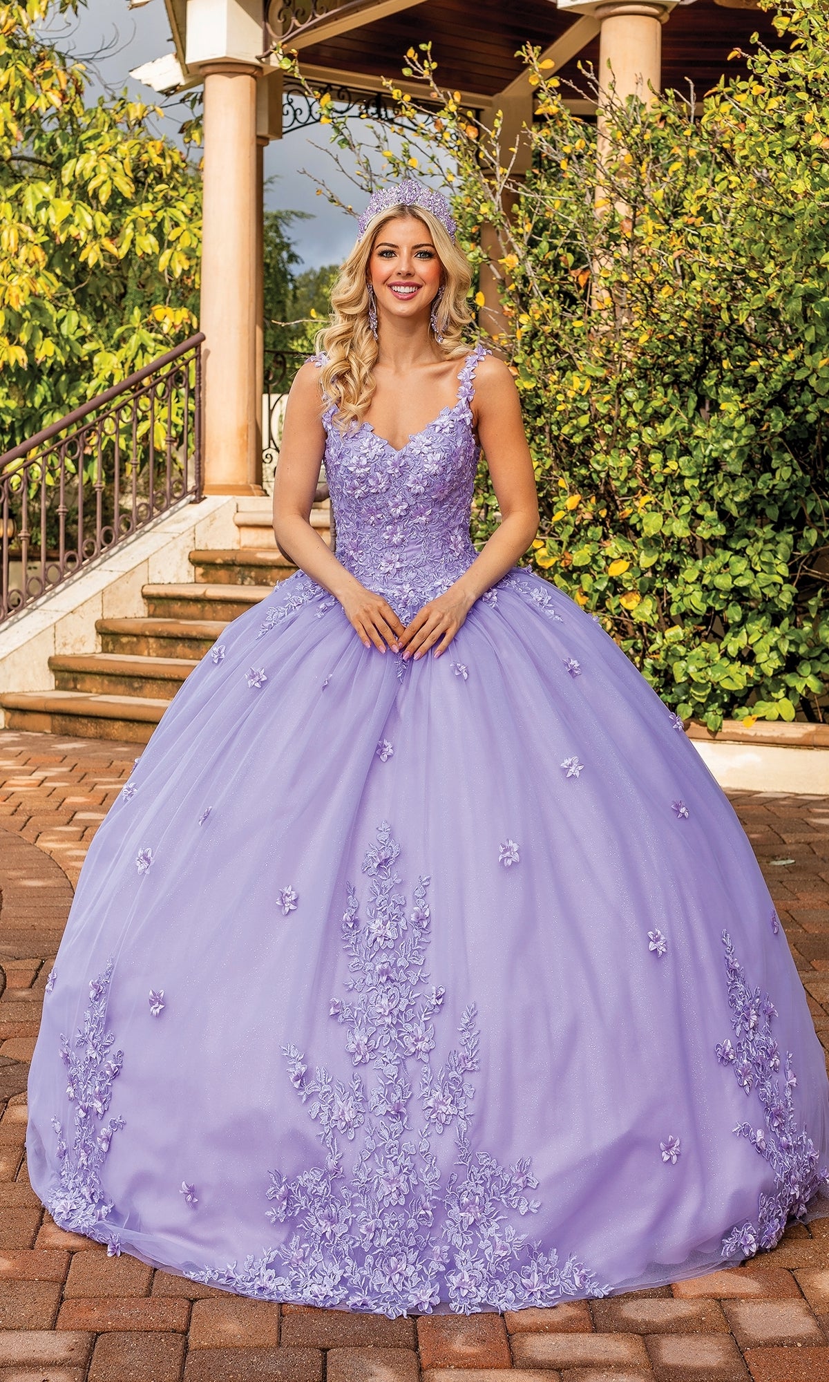A woman stands outdoors in the Quinceanera Dress 1898 by Dancing Queen, a lavender a-line ball gown with floral embellishments, posing before stairs and lush greenery.