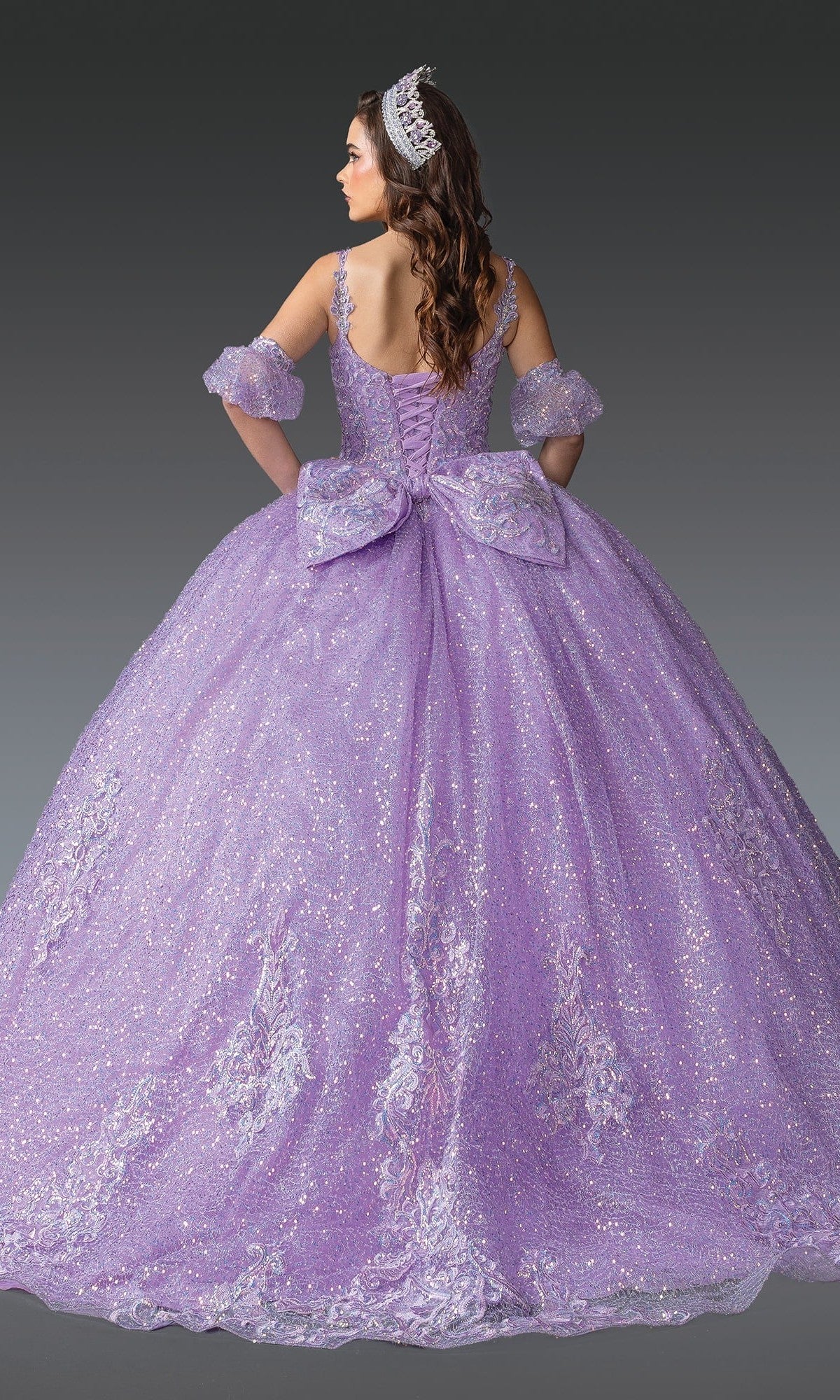 A woman wearing the Quinceanera Dress 1907 by Dancing Queen—featuring sparkling lavender glitter, delicate lace details, a large bow, detachable off-shoulder sleeves, and a tiara—is seen from the back against a plain gray background, showcasing the elegant silhouette of this stunning gown.