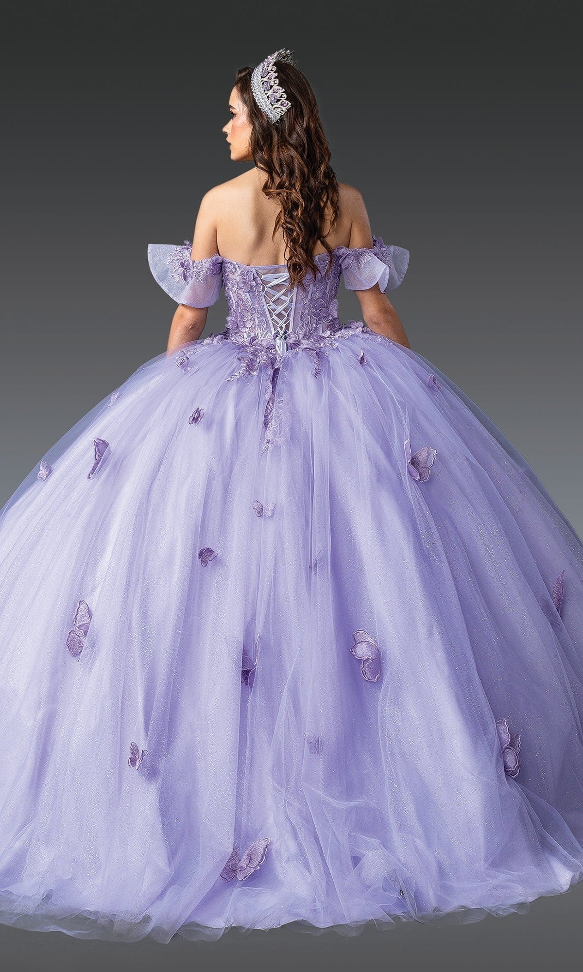 A woman stands against a plain gray background wearing the Quinceanera Dress 1911 by Dancing Queen—an off-shoulder lavender ball gown adorned with delicate butterfly appliques, featuring a dramatic ballgown skirt, a lace-up back, and paired with a silver tiara.